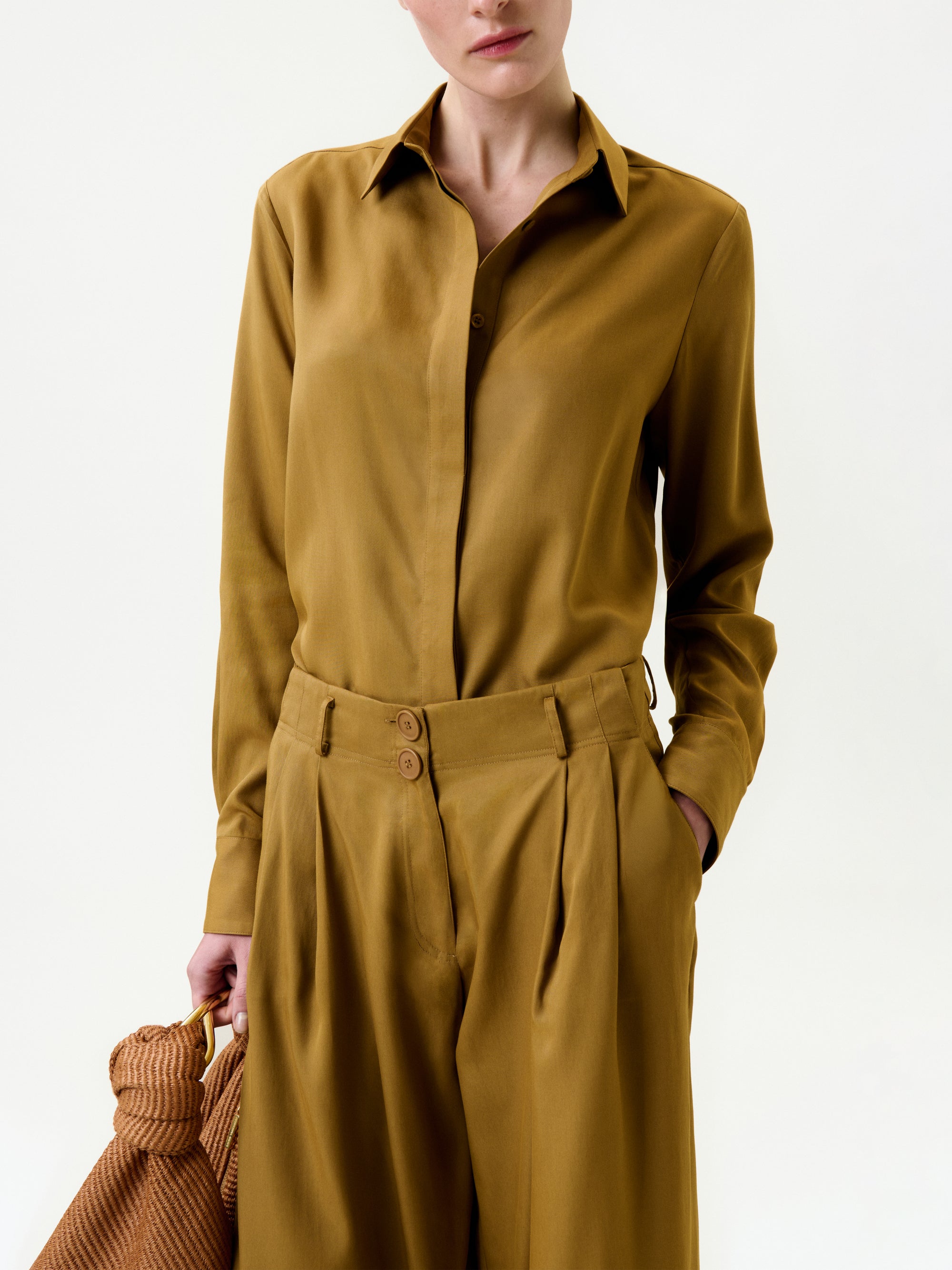 A person in the Mazz Blouse, a mustard yellow collared shirt made from 100% Lyocell Italian fabric, stands with one hand in their pocket holding a textured brown bag. Made in Portugal, shown against a plain light background.