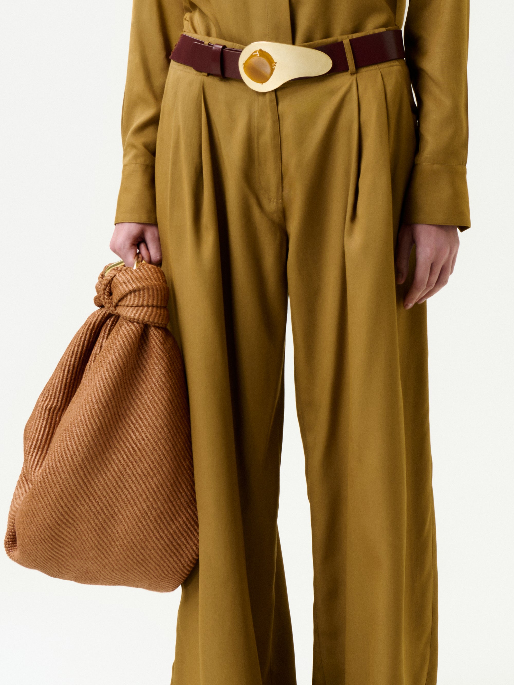 A person wears the Opal Pant in mustard, made from fine Italian fabric with pleats, paired with a burgundy belt with a large round buckle, and holds a light brown knotted textured bag in their left hand.