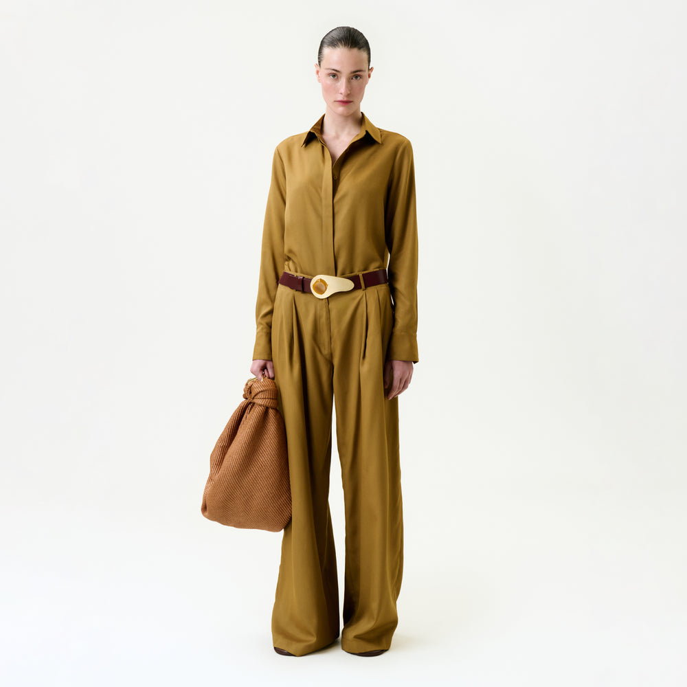 A person stands against a plain white background, wearing the Opal Pant with a mustard brown Italian-fabric blouse, holding a textured brown bag, and accessorized with a wide belt featuring a round buckle.