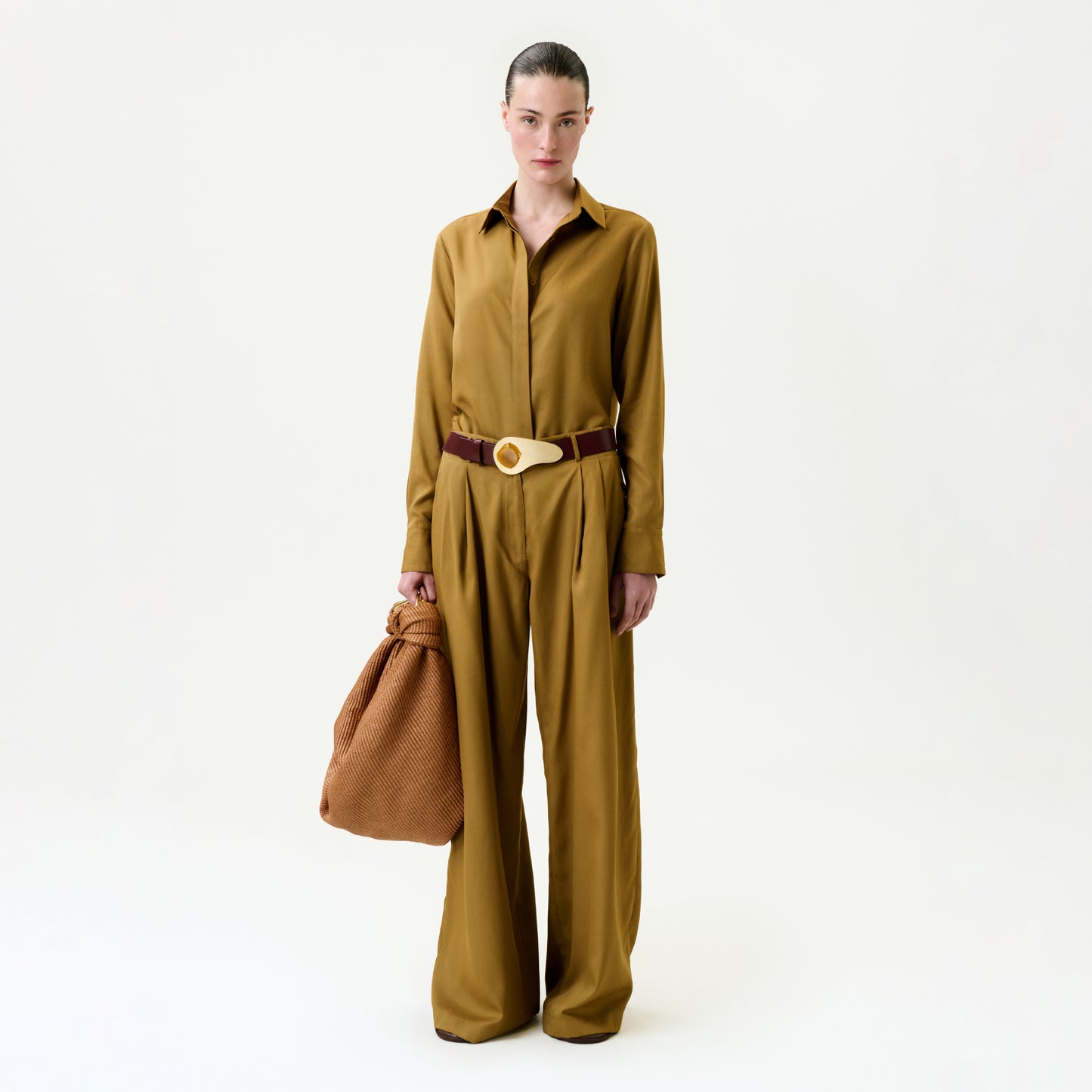 A person stands against a plain white background, wearing the Opal Pant with a mustard brown Italian-fabric blouse, holding a textured brown bag, and accessorized with a wide belt featuring a round buckle.