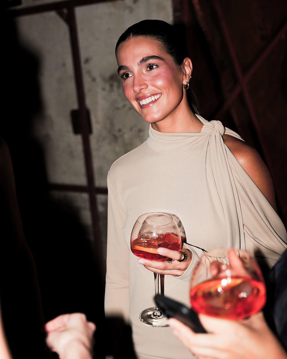 A woman smiling and holding a cocktail glass in a dimly lit social setting, wearing a beige draped high-neck top.