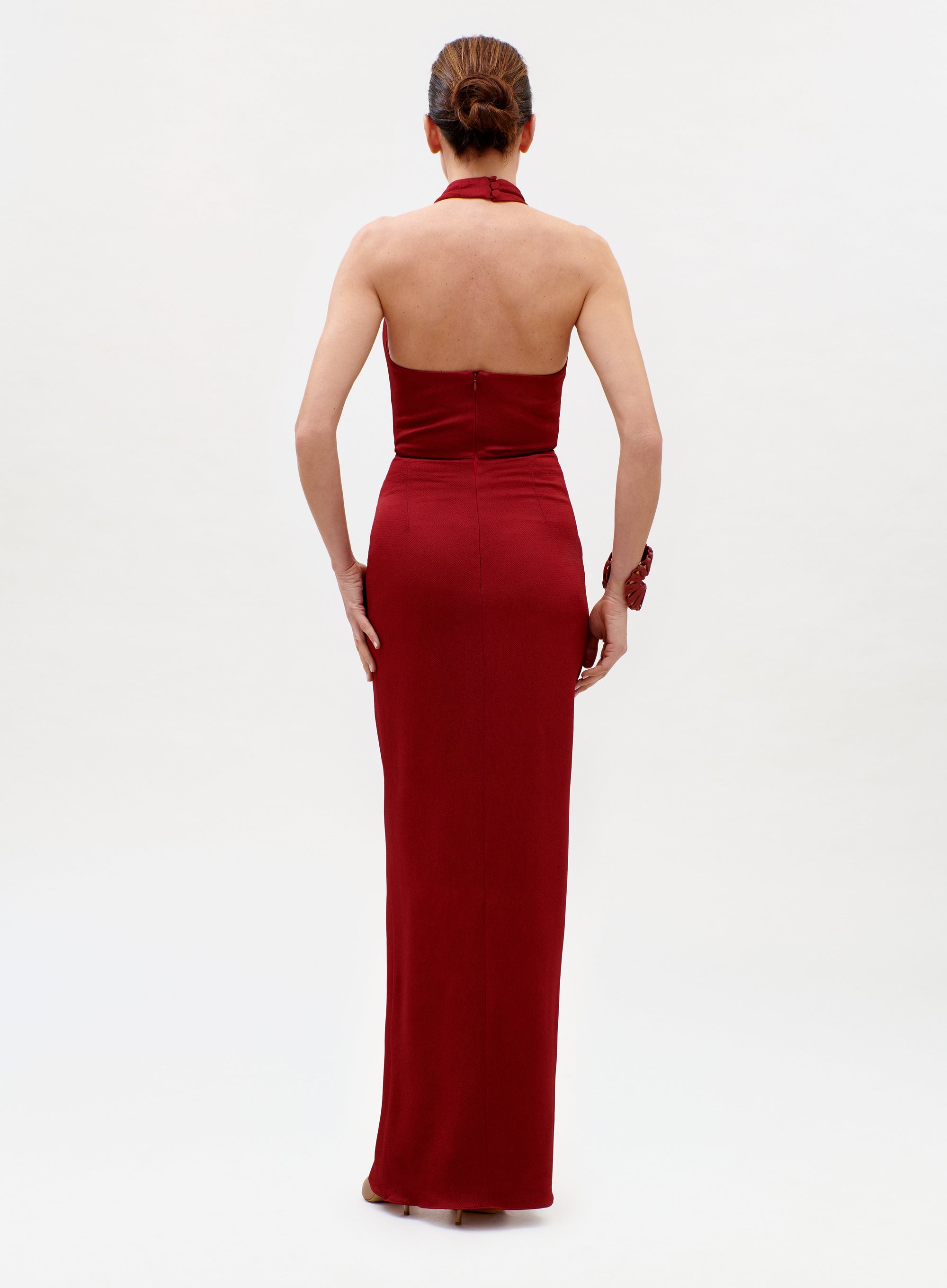 A woman with her hair in a bun stands with her back to the camera, wearing the long, sleeveless Davinia Dress in red against a plain white background.
