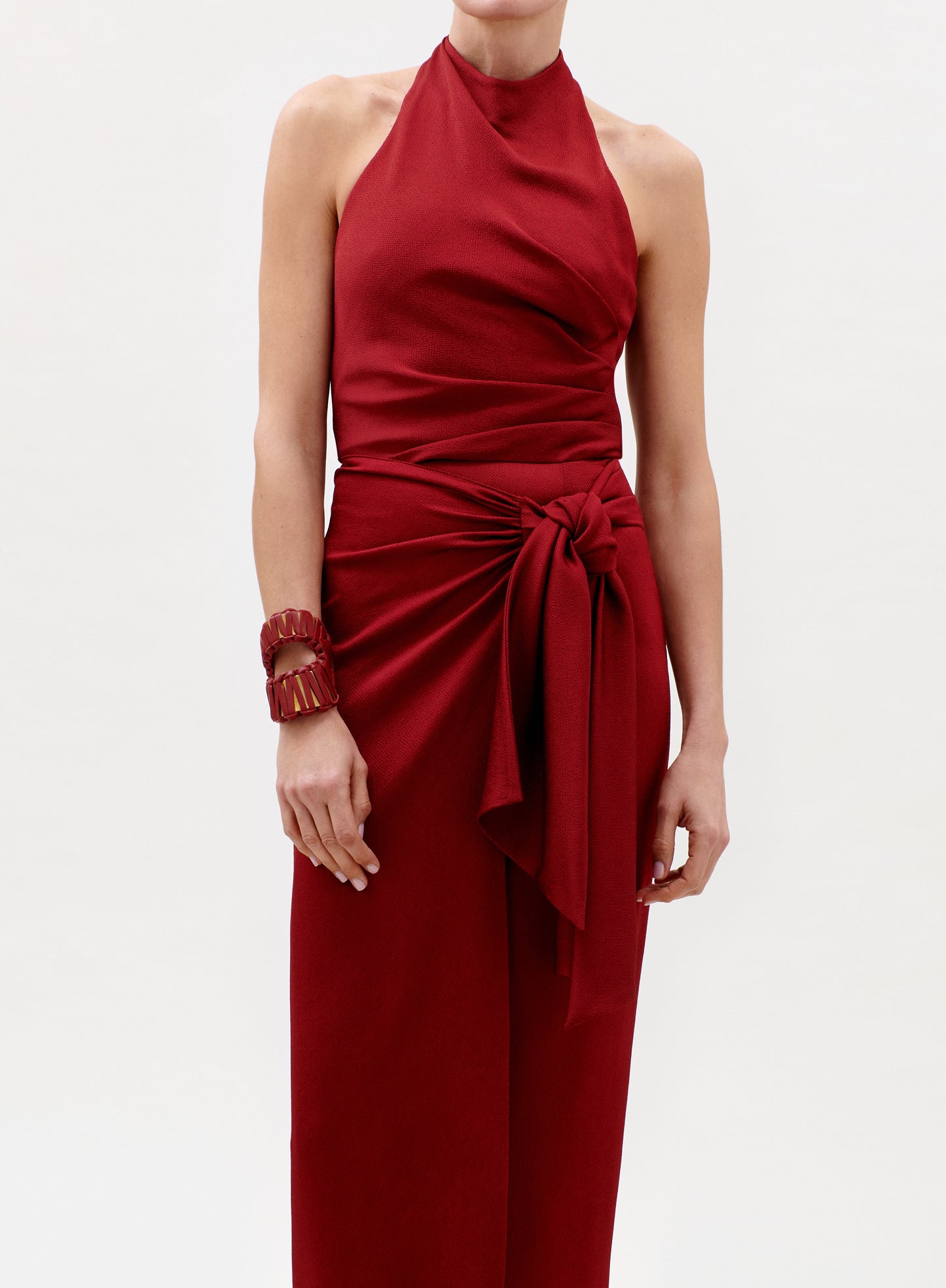A person wearing the Davinia Dress—a sleeveless, high-neck red dress with a large bow at the waist—and a matching red bracelet, standing against a plain light background.
