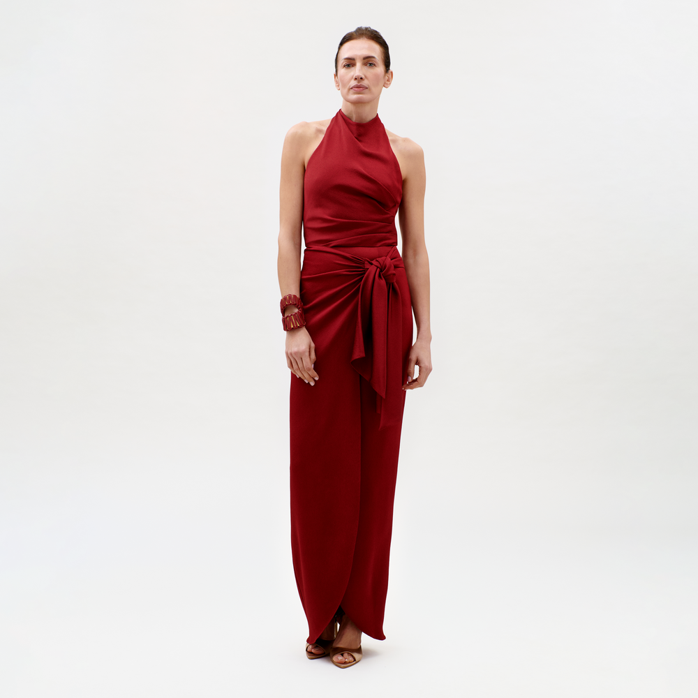 A woman poses against a plain light background in the Davinia Dress—a sleeveless, halter-neck, floor-length red dress with a side waist tie—paired with brown heeled sandals and a matching red bracelet.