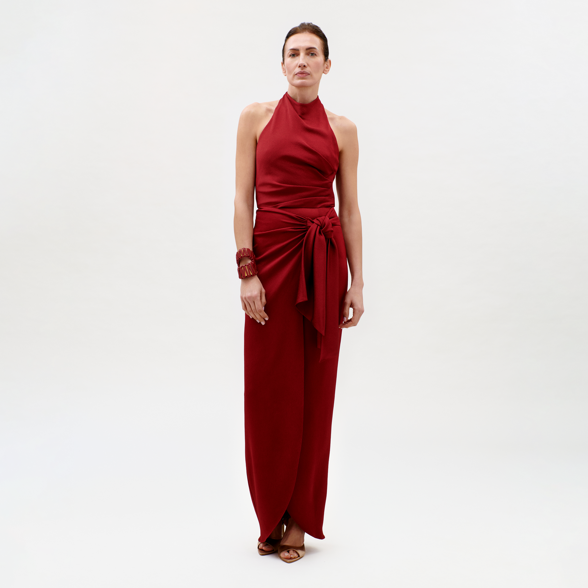 A woman poses against a plain light background in the Davinia Dress—a sleeveless, halter-neck, floor-length red dress with a side waist tie—paired with brown heeled sandals and a matching red bracelet.