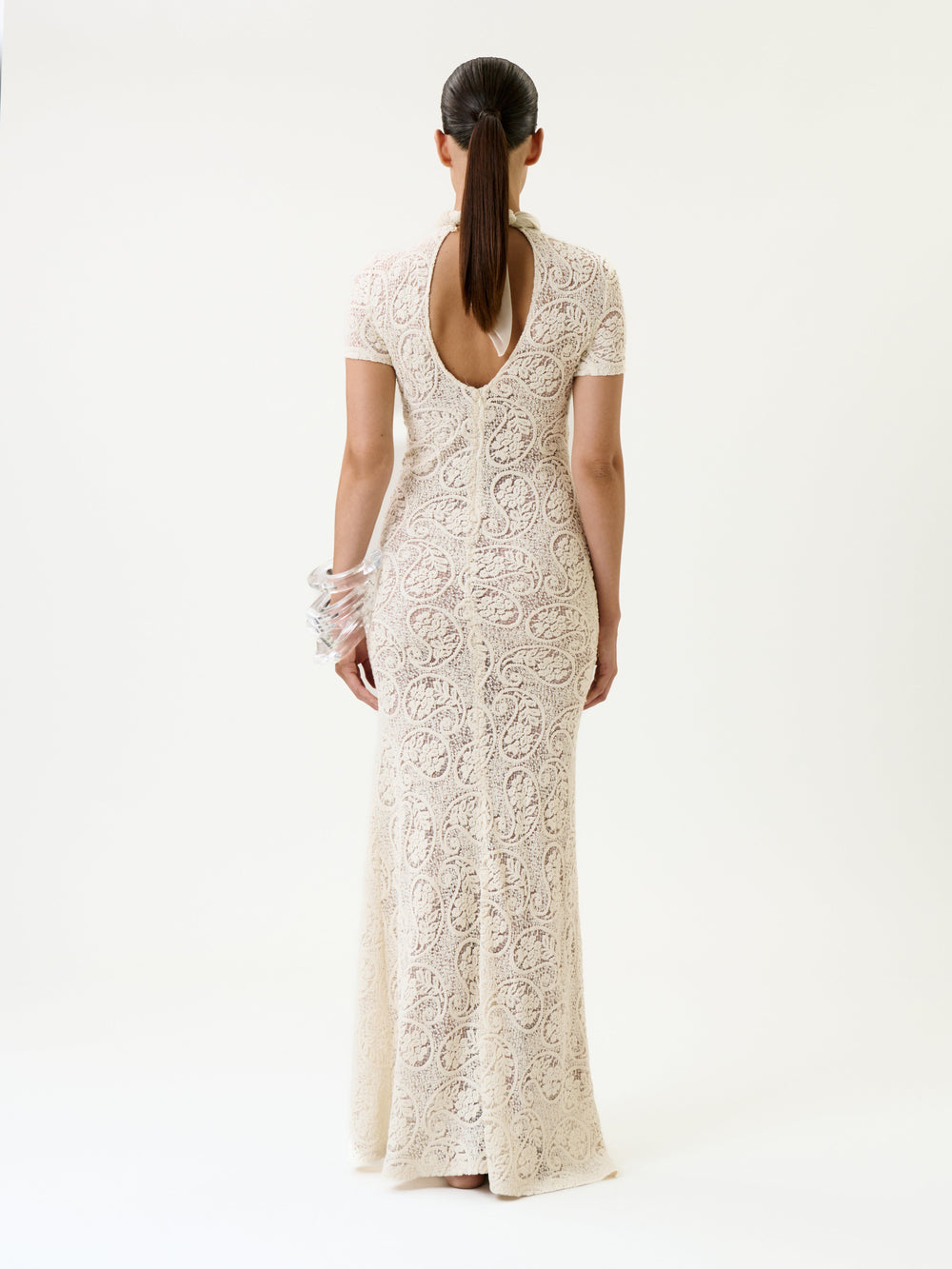 A woman stands facing away, wearing the Tini Dress—a long, fitted cream lace design featuring a keyhole back and short sleeves. Her hair is tied back, and she wears translucent bangles on her left wrist against a plain background.