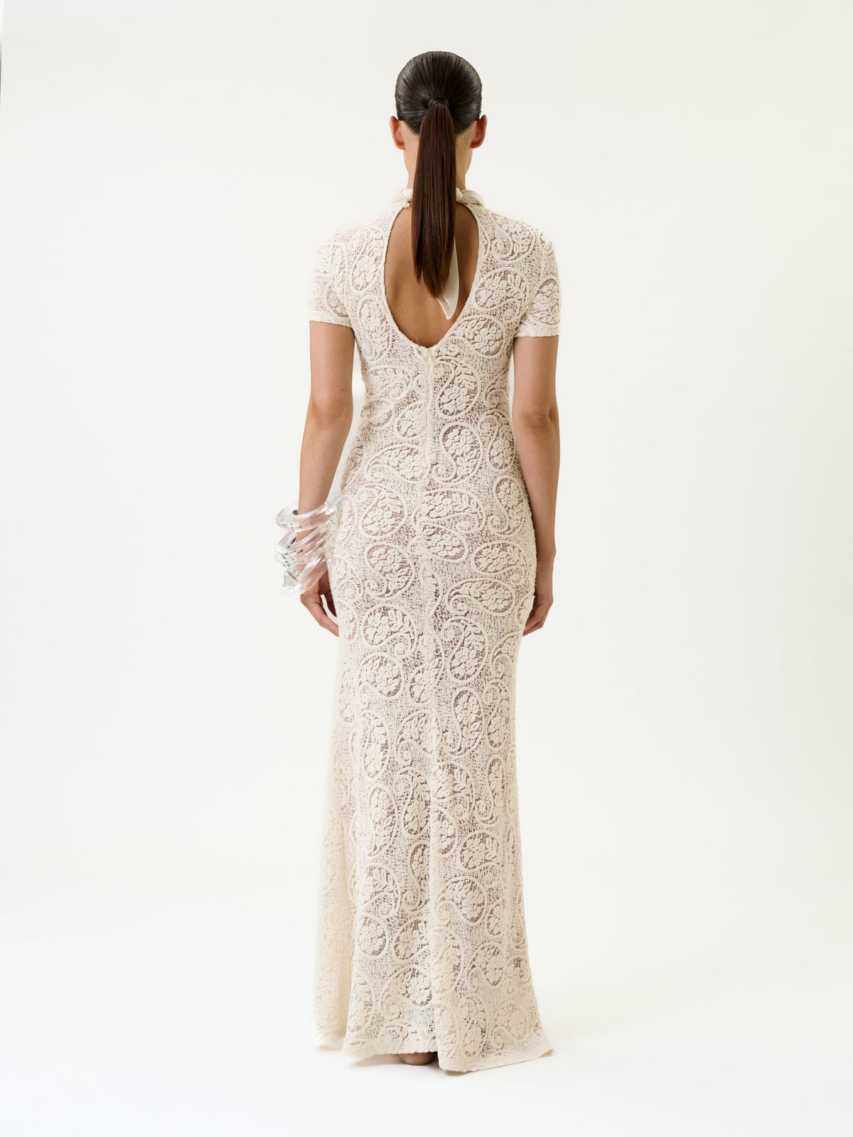 A woman stands facing away, wearing the Tini Dress—a long, fitted cream lace design featuring a keyhole back and short sleeves. Her hair is tied back, and she wears translucent bangles on her left wrist against a plain background.