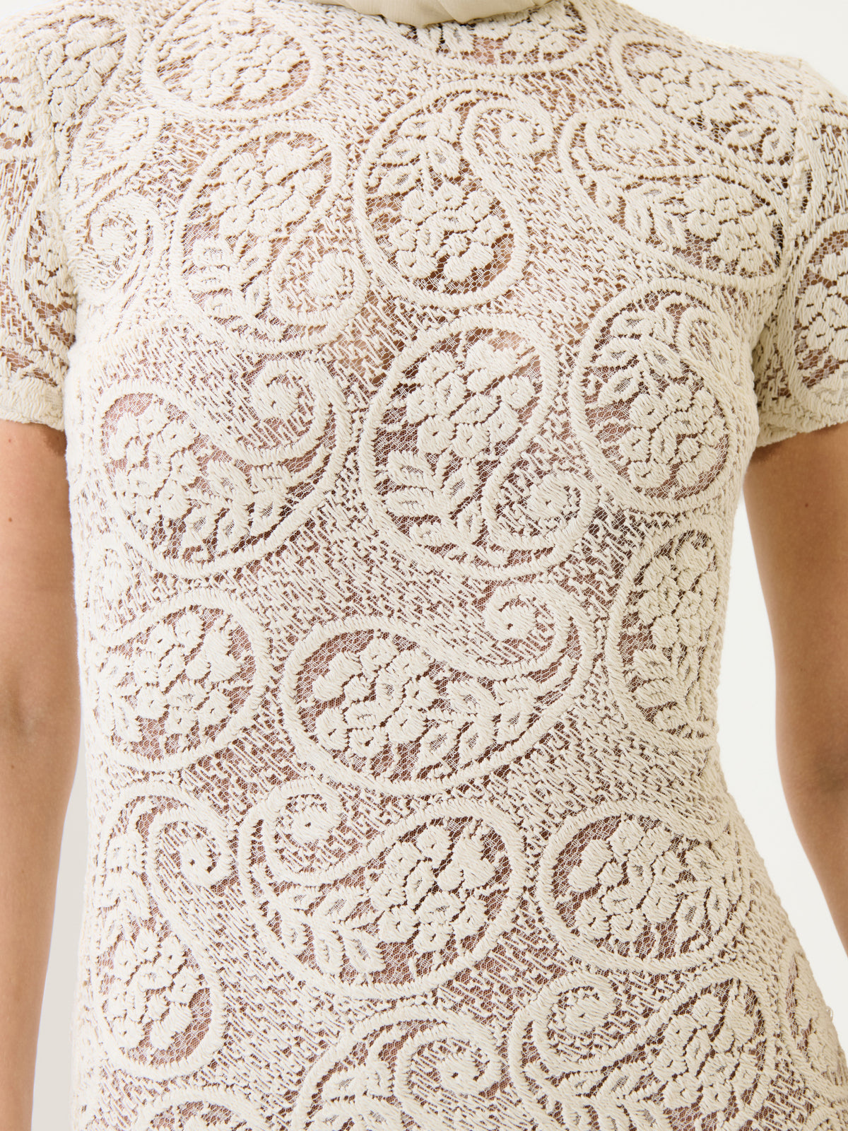 Close-up of a person wearing the Tini Dress—a fitted, cream-colored lace dress featuring a sheer finish and an all-over paisley floral pattern. The intricate lace design covers the entire visible area of the dress.