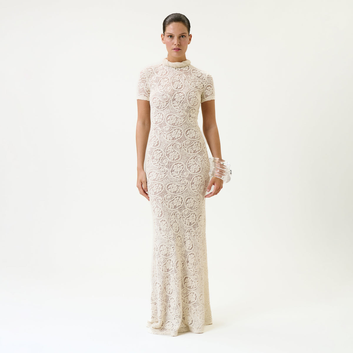 A woman stands against a plain white background wearing the floor-length, short-sleeved Tini Dress in cream lace with a high neckline and a clear bangle on her wrist.