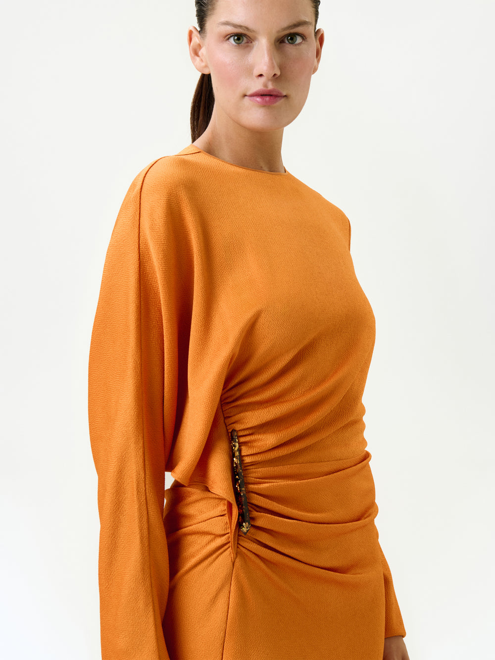 A woman with straight brown hair pulled back wears the Shiloh Dress, an orange fitted midi crafted from Italian fabric with ruched waist detailing and embellishments, standing against a plain white background.
