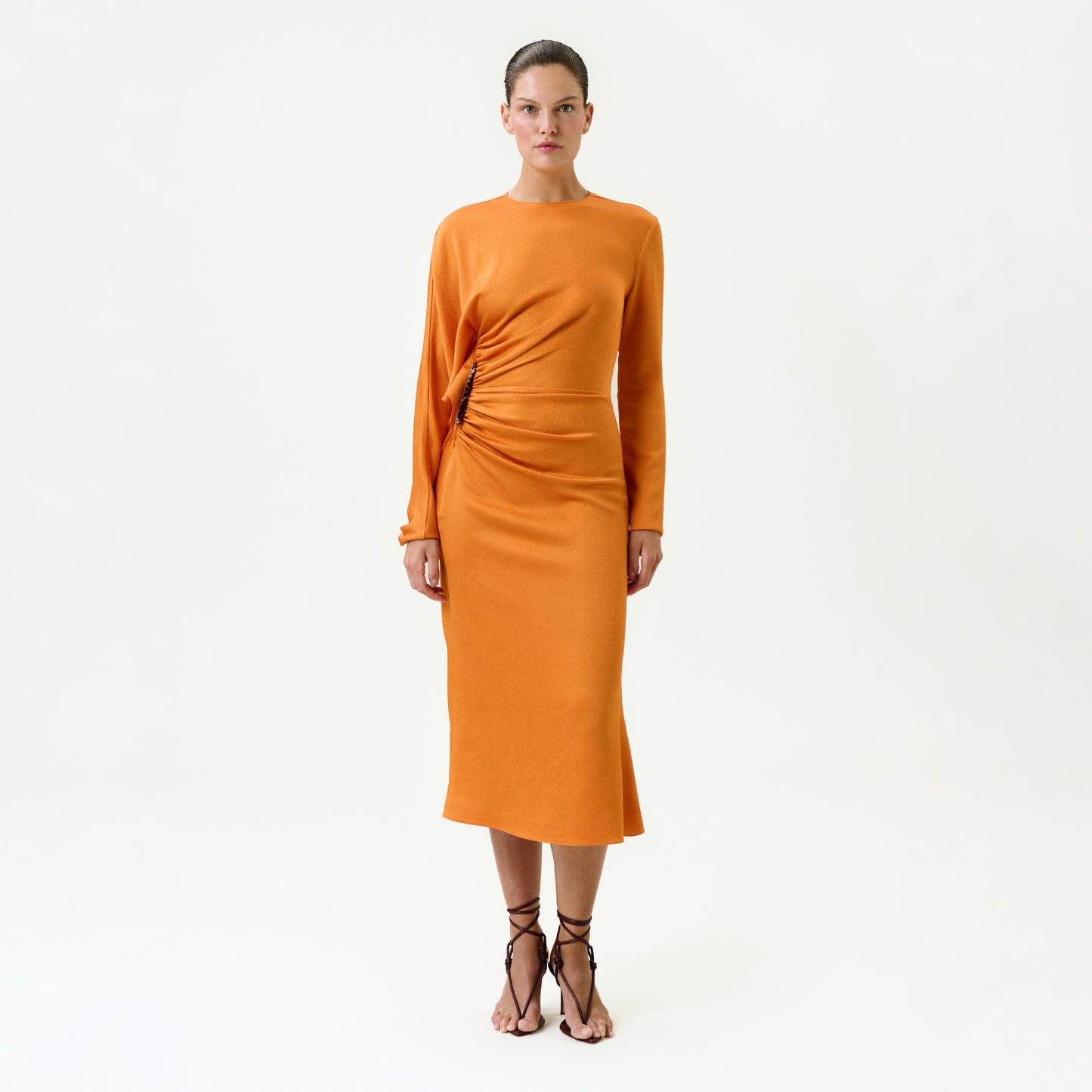 A woman faces forward against a white background, wearing the Shiloh Dress—a long-sleeved, fitted orange midi dress made from Italian fabric with a gathered waist—and strappy black sandals. She has a neutral expression.