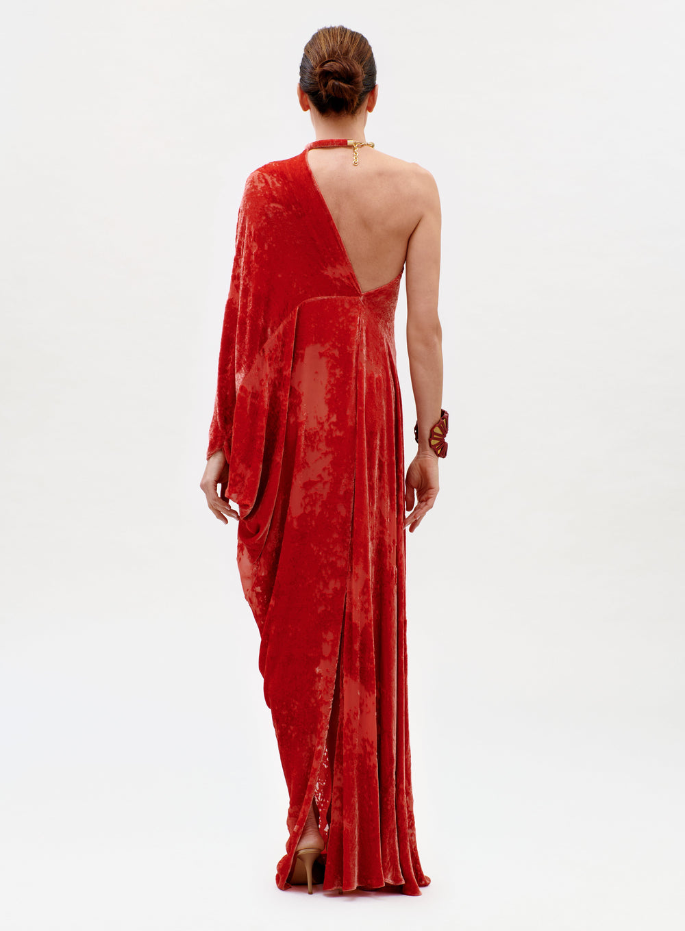 A person stands with their back turned, wearing the Erica Dress—a textured, one-shoulder red gown with a draped sleeve and long skirt. Their hair is styled in a low bun, paired with gold necklace and bracelet accessories.