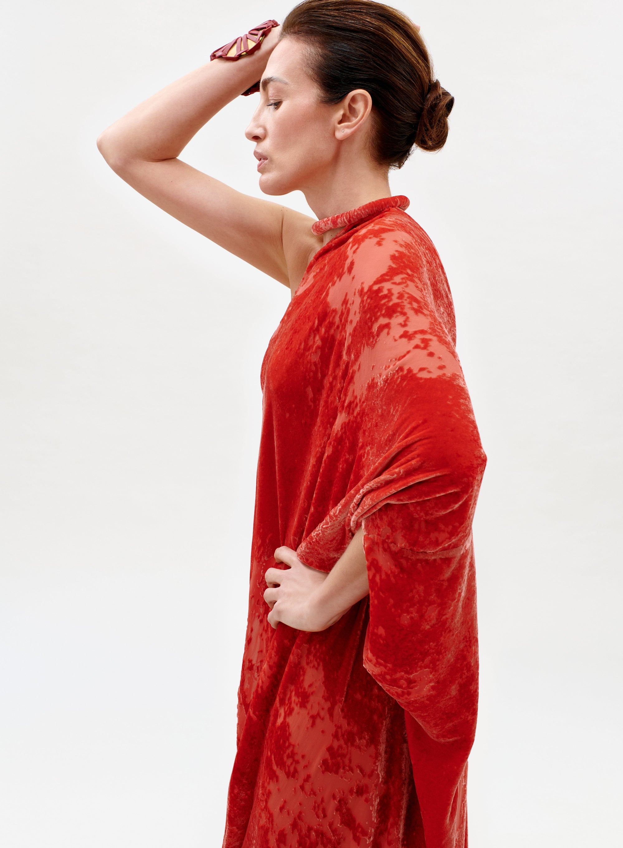 A woman in the Erica Dress, featuring a textured red fabric, stands in profile against a plain background with one arm raised to her forehead and her hair styled in a neat bun.