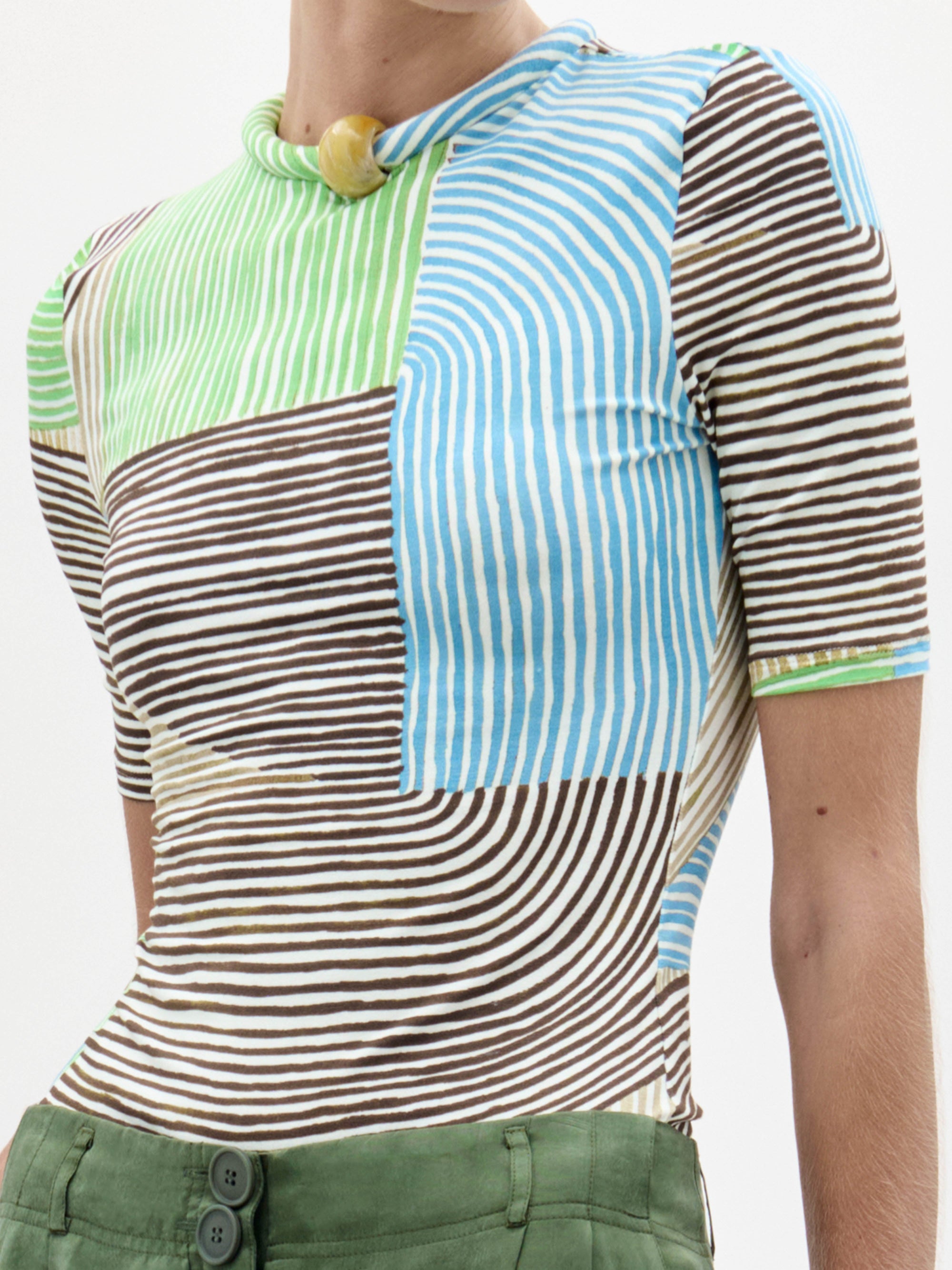 A woman stands against a plain background wearing the Varinia Tshirt with stripes and loose-fitting green trousers, holding a small yellow bag.