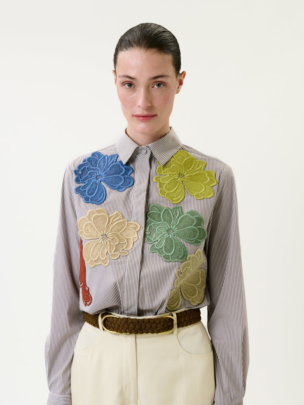 A woman with slicked-back hair wears the Warda Shirt, a striped top adorned with large, colorful floral appliqués, paired with high-waisted cream trousers and a braided brown belt, standing against a plain light background.
