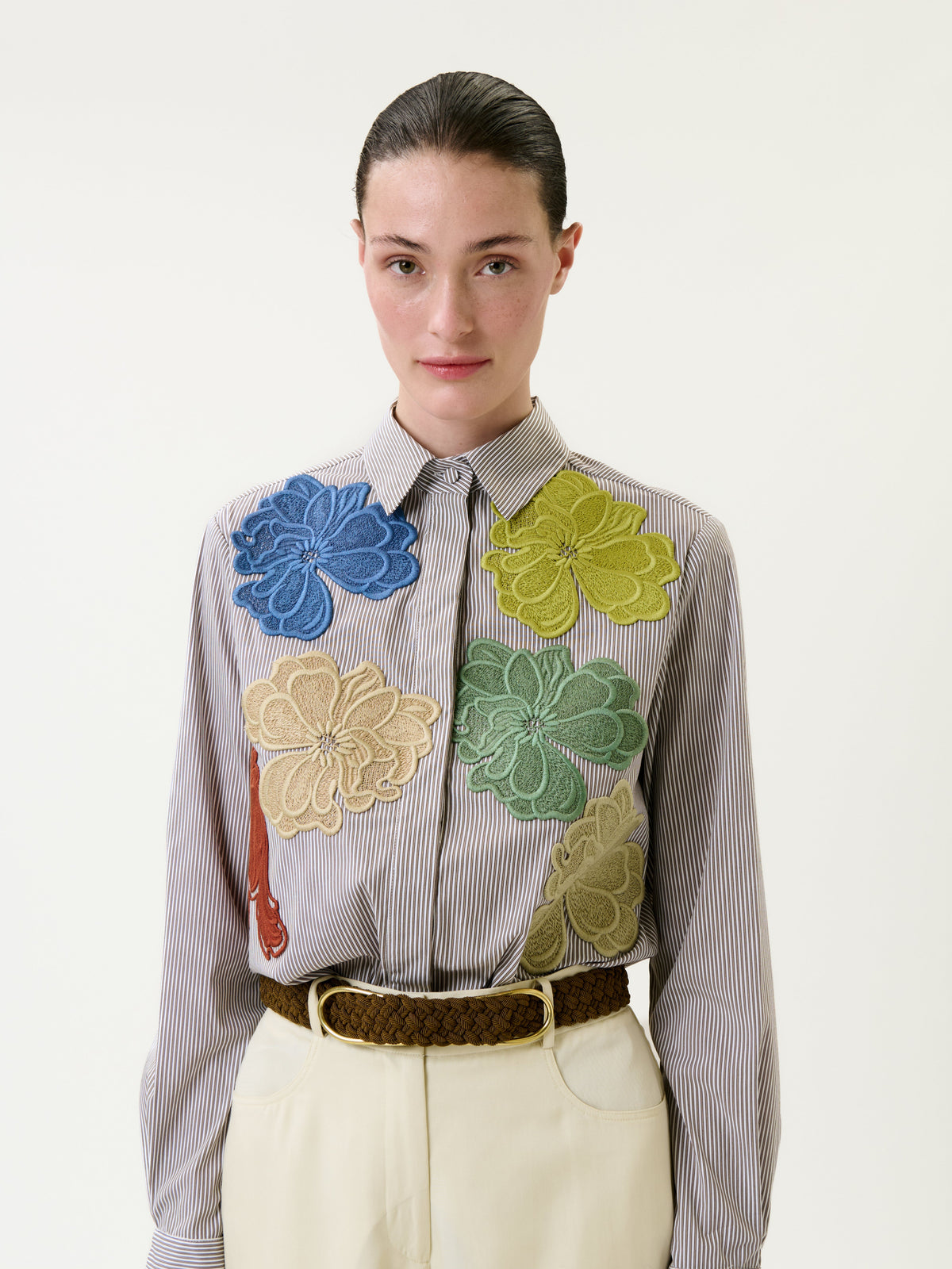 A woman with slicked-back hair wears the Warda Shirt, a striped top adorned with large, colorful floral appliqués, paired with high-waisted cream trousers and a braided brown belt, standing against a plain light background.