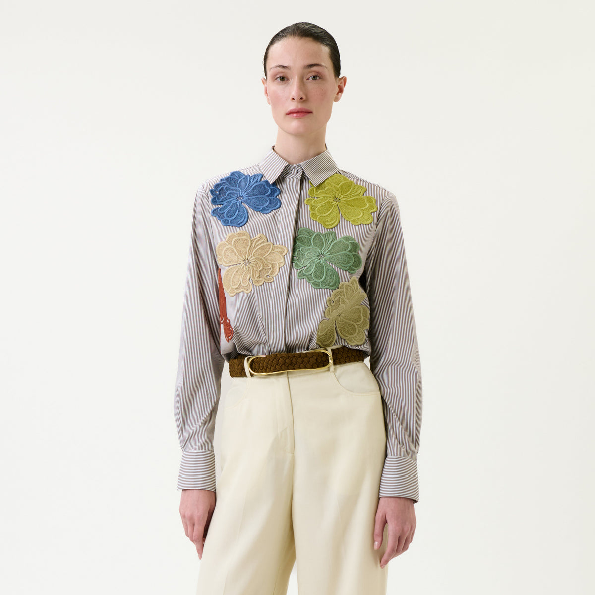 A person stands against a white background wearing the Warda Shirt—a striped top adorned with large, colorful floral appliqués—paired with cream high-waisted pants and a brown woven belt.