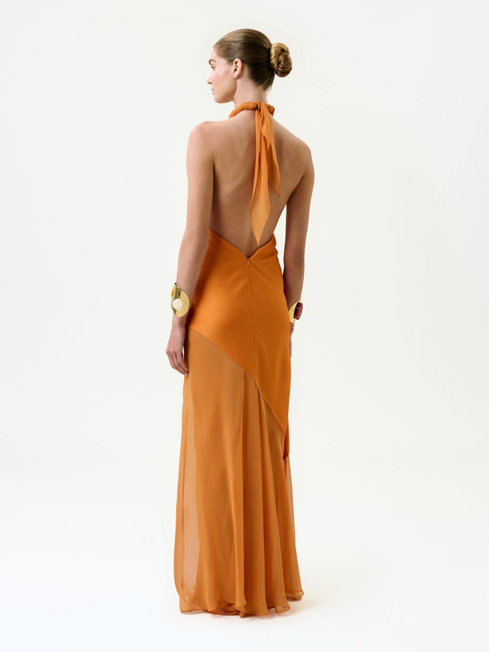 A person with a neat bun stands against a plain white background, wearing the Keisha Dress—a backless, halter-neck orange silk maxi dress with a sheer flowing skirt—styled with chunky gold bracelets on both wrists.