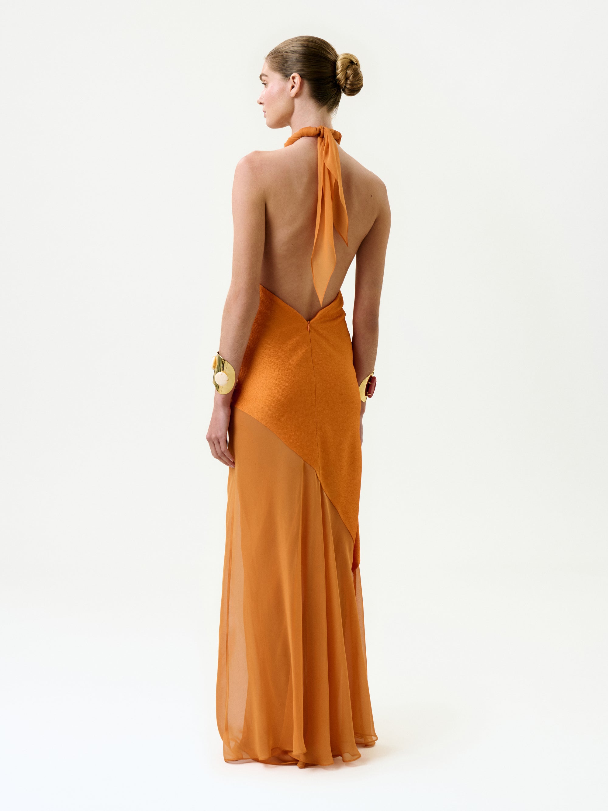 A person with a neat bun stands against a plain white background, wearing the Keisha Dress—a backless, halter-neck orange silk maxi dress with a sheer flowing skirt—styled with chunky gold bracelets on both wrists.