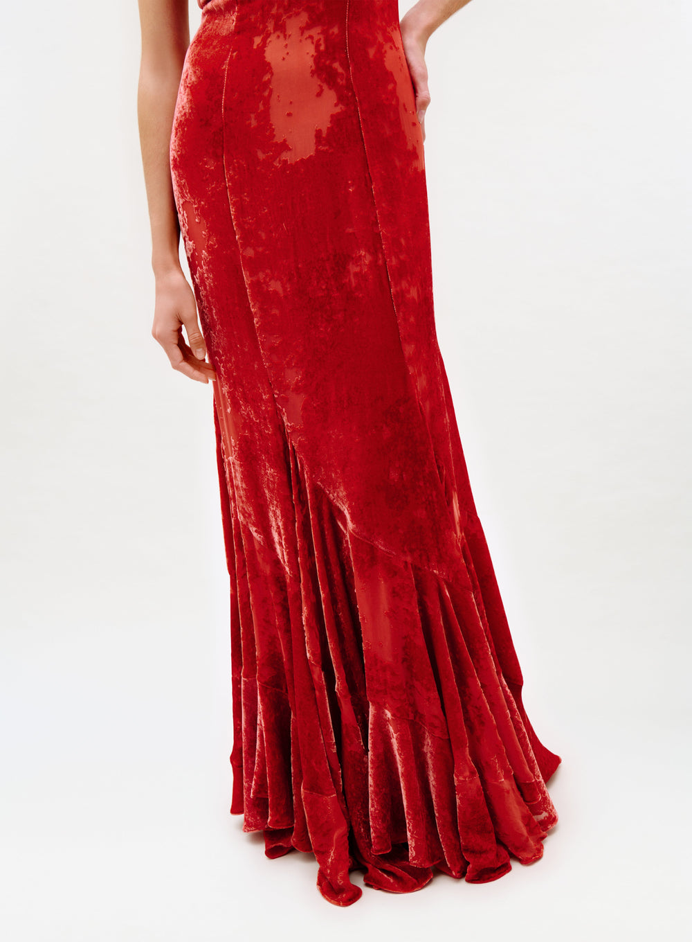 Wearing the Curitiba Dress—a long, fitted red velvet dress with a flared hem and shimmering textured finish—someone stands against a plain white background, showing only the lower body.