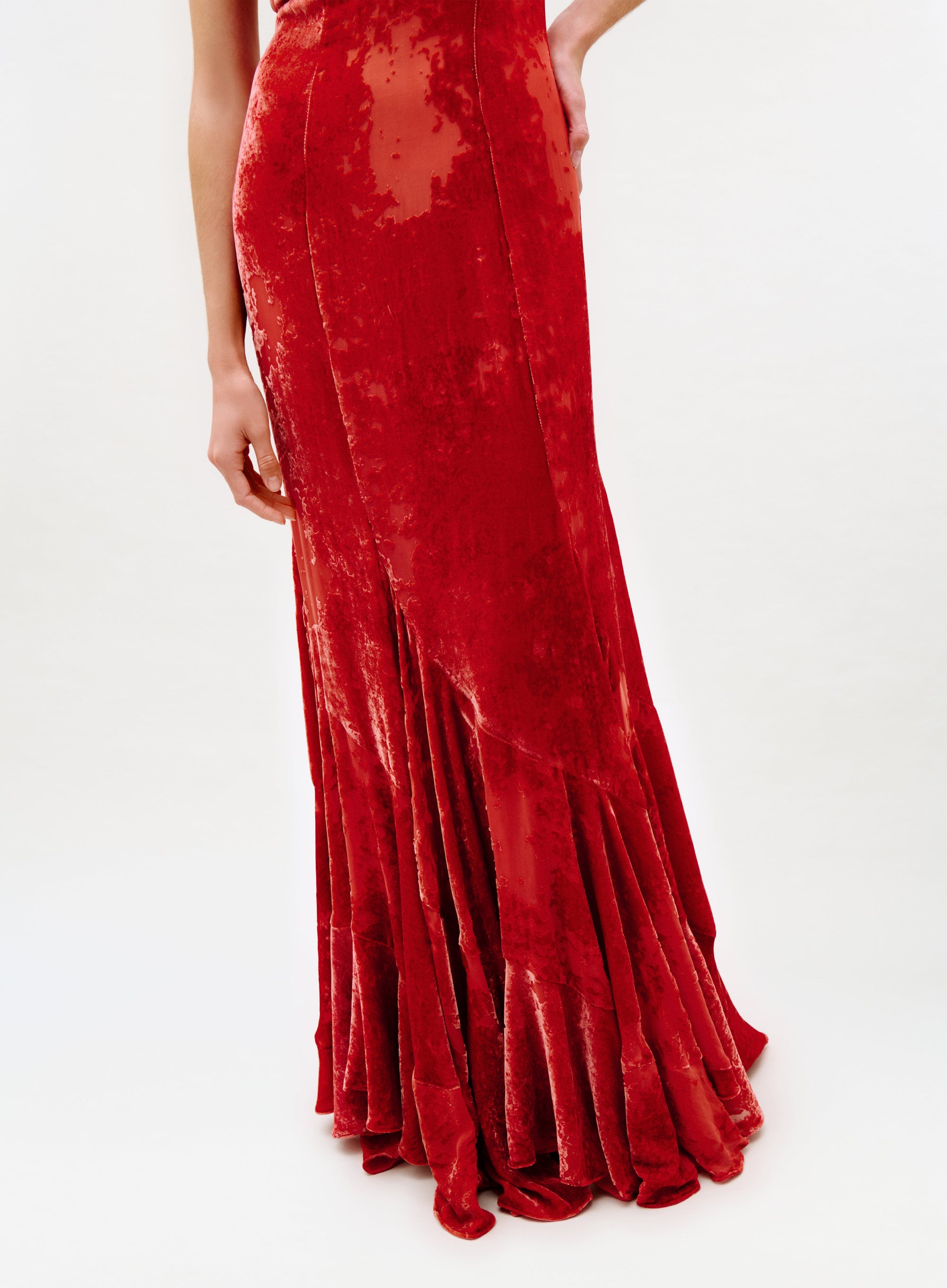 Wearing the Curitiba Dress—a long, fitted red velvet dress with a flared hem and shimmering textured finish—someone stands against a plain white background, showing only the lower body.