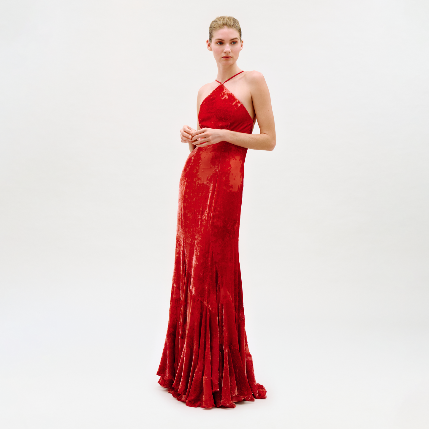 A person stands against a plain white background wearing the Curitiba Dress, a floor-length, sleeveless red velvet gown with thin crisscross straps and a fitted silhouette.