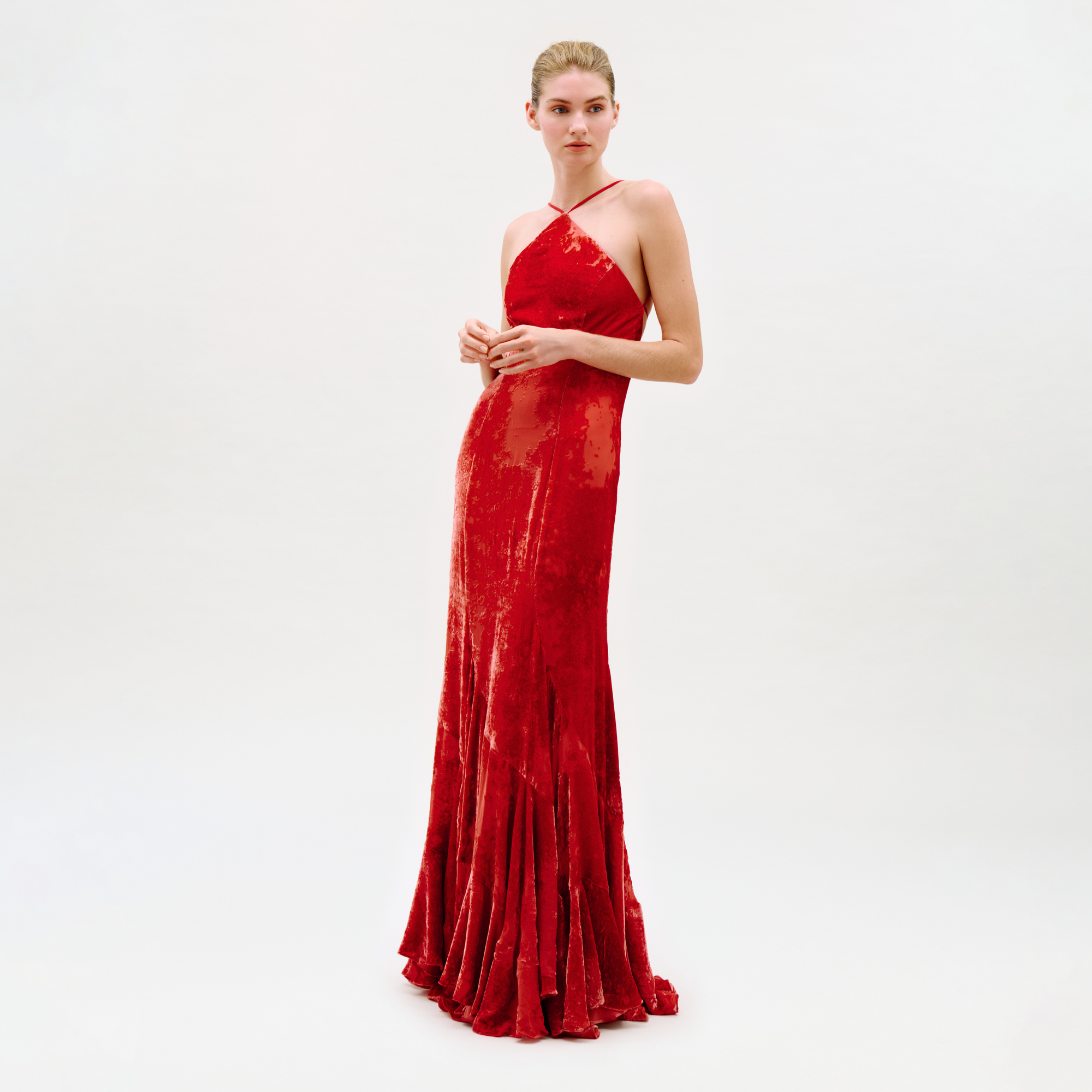 A person stands against a plain white background wearing the Curitiba Dress, a floor-length, sleeveless red velvet gown with thin crisscross straps and a fitted silhouette.