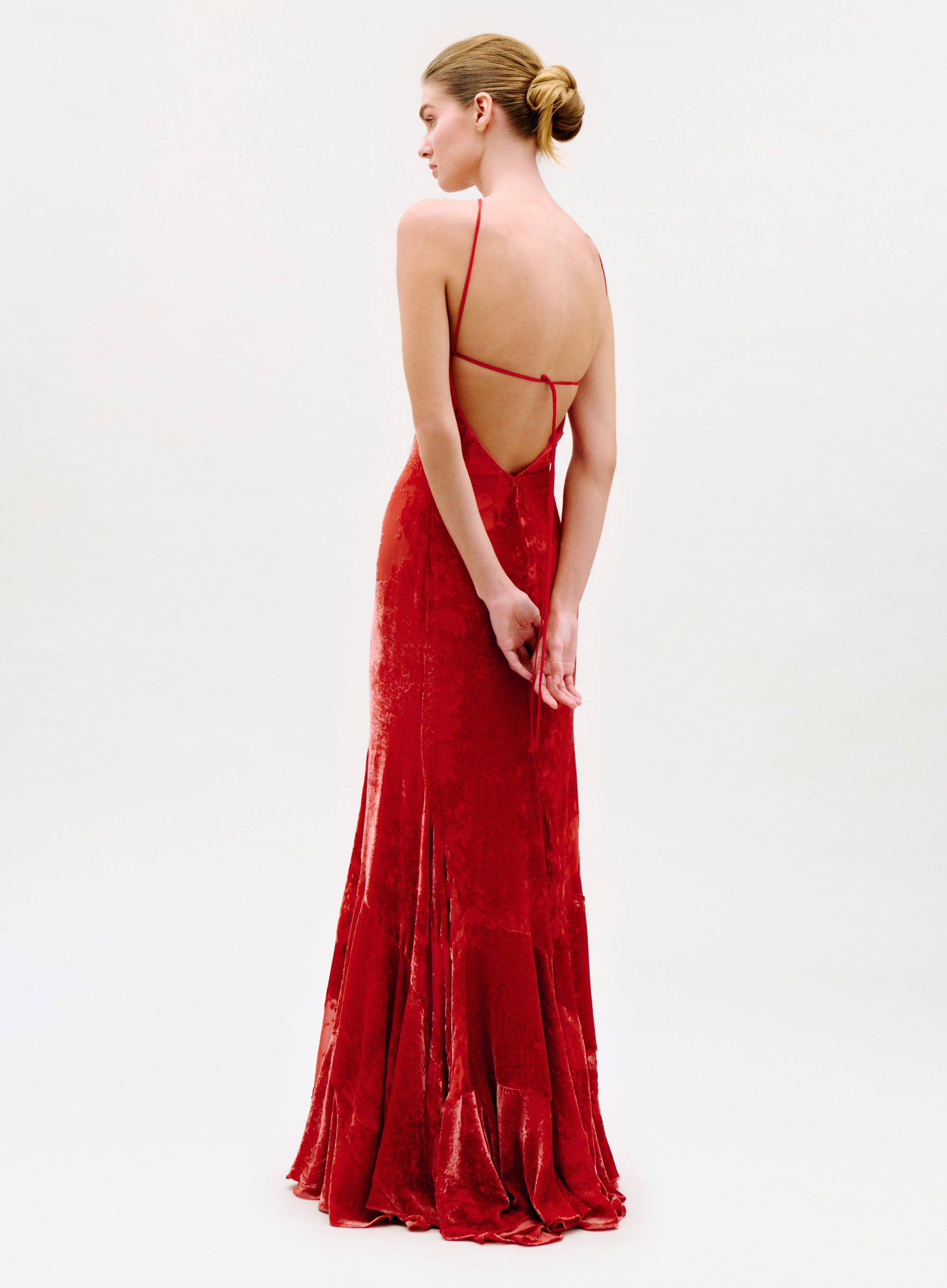 A woman in the floor-length Curitiba Dress—with thin straps, a low-cut open back, and red velvet fabric—stands against a plain light background, looking to the side with her hands clasped behind her.