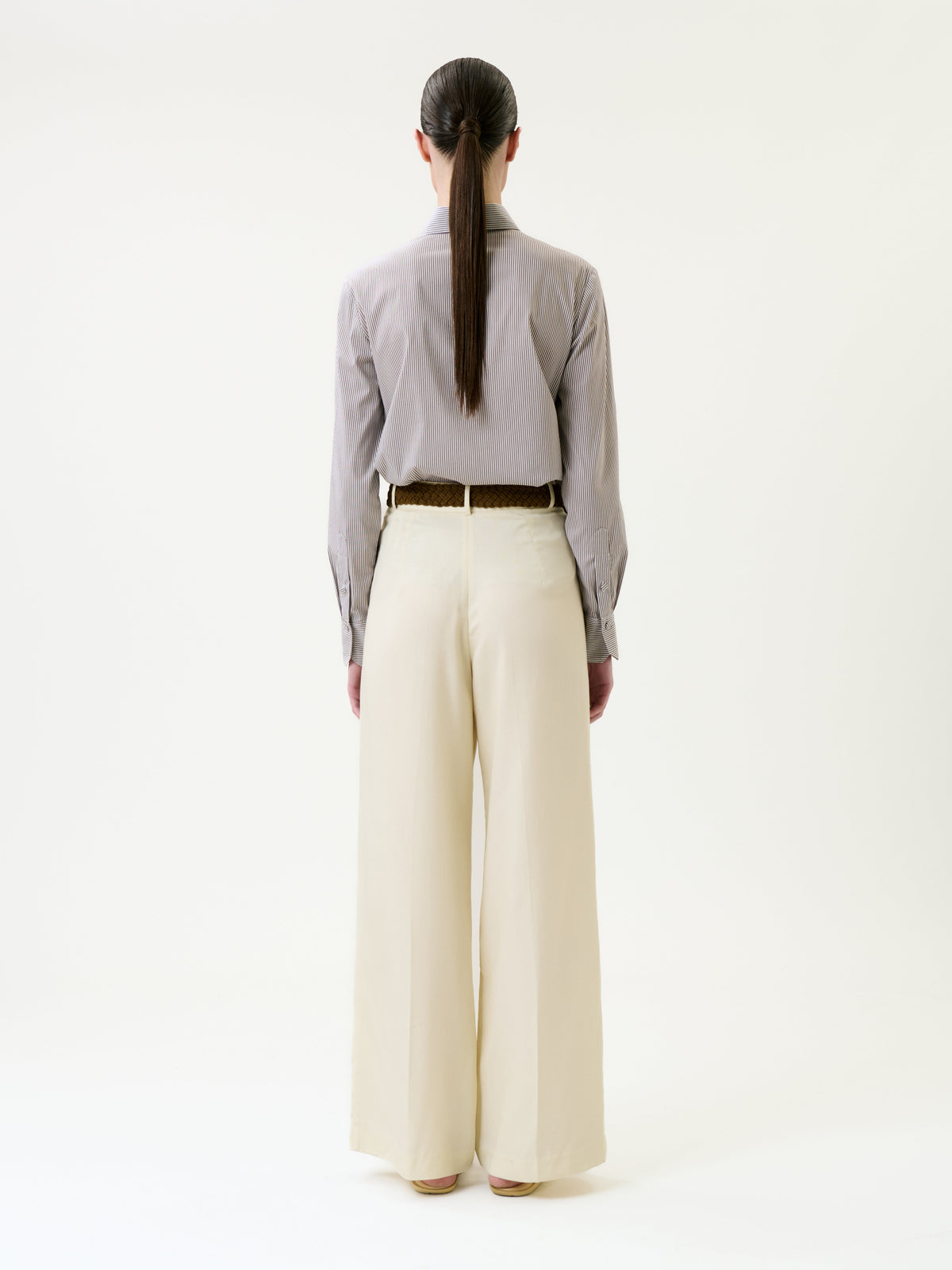 A person with long brown hair in a ponytail is standing facing away, wearing a long-sleeved gray shirt tucked into the high-waisted, wide-leg Emine Pant in cream and a brown belt, against a plain white background.