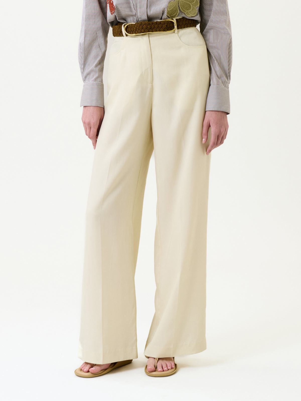 Wearing the Emine Pant in cream, styled with a braided brown belt, a tucked-in striped shirt featuring an embroidered patch, and beige sandals, photographed against a plain white background.