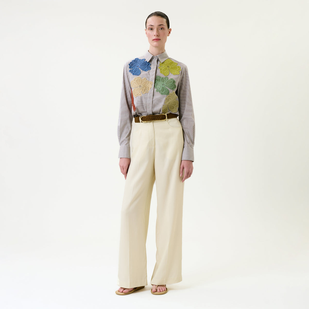 A person stands against a white background wearing a striped shirt with large colorful flower appliqués, the Emine Pant in beige wide-leg style, a brown belt, and sandals.