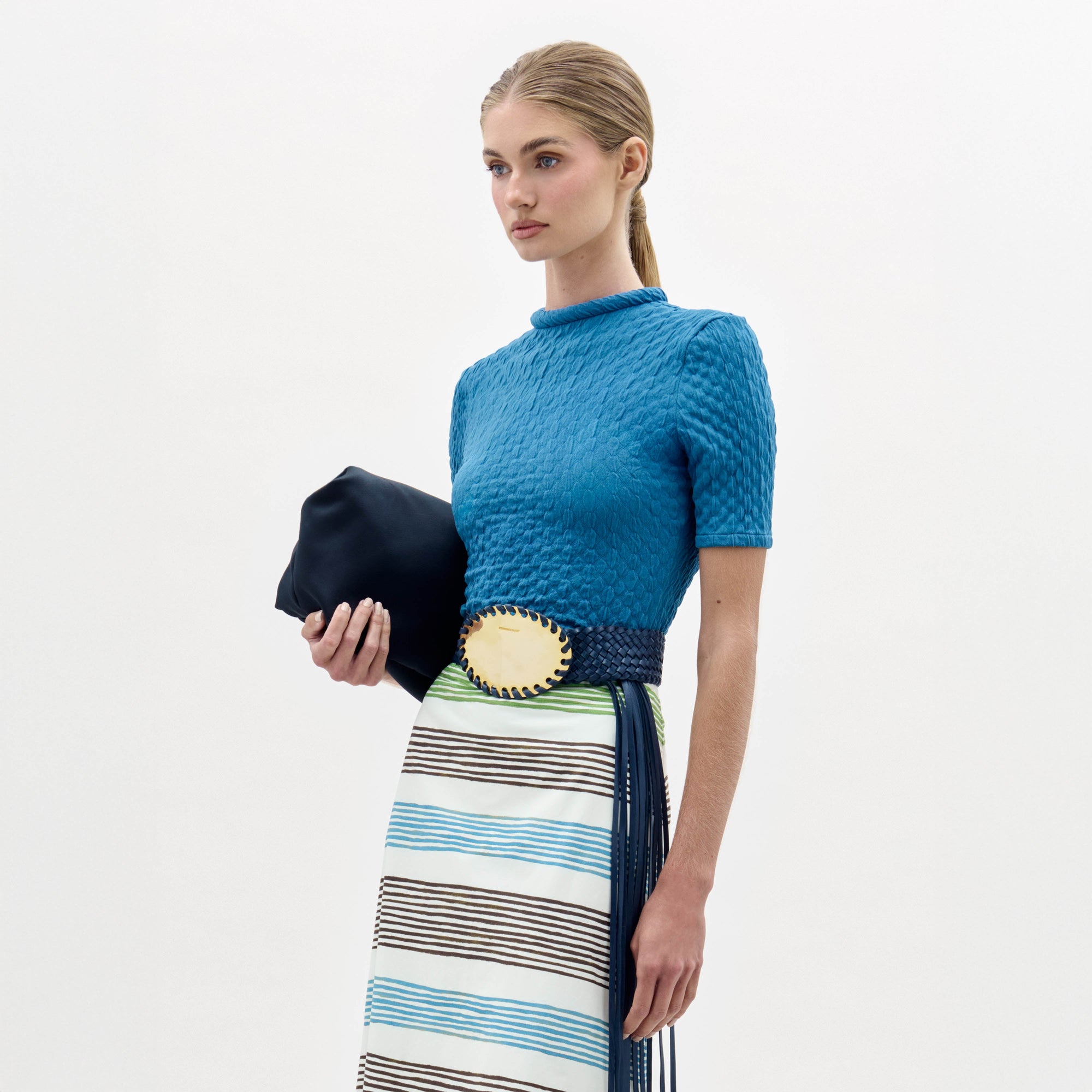 A woman in a blue Venecia Tshirt and striped skirt holds a black clutch. Her straight hair is pulled back, and she wears a large oval belt buckle.