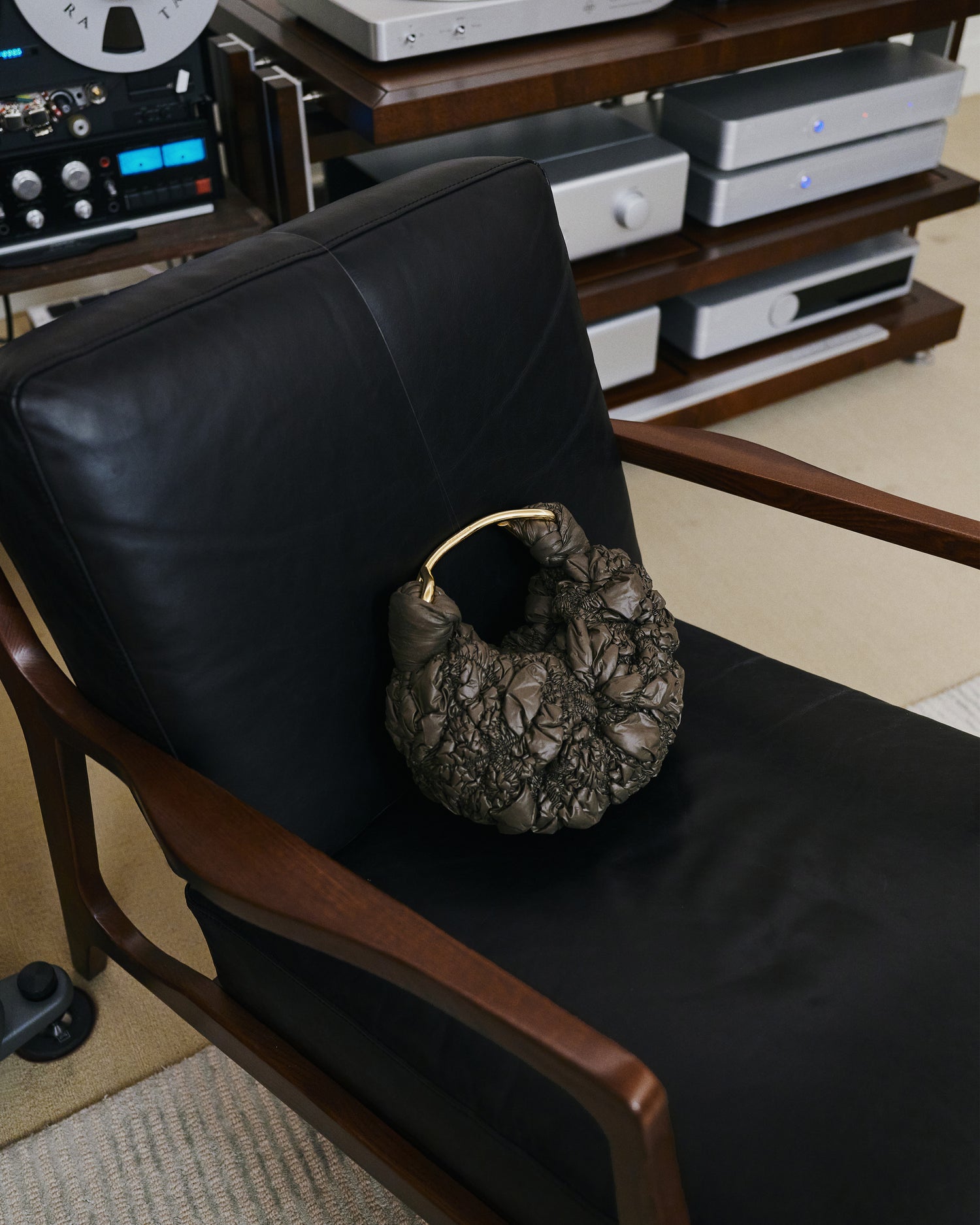 A textured metallic bronze crumpled-paper style clutch bag with a gold-tone handle resting on a black leather armchair in a room with audio equipment in the background.