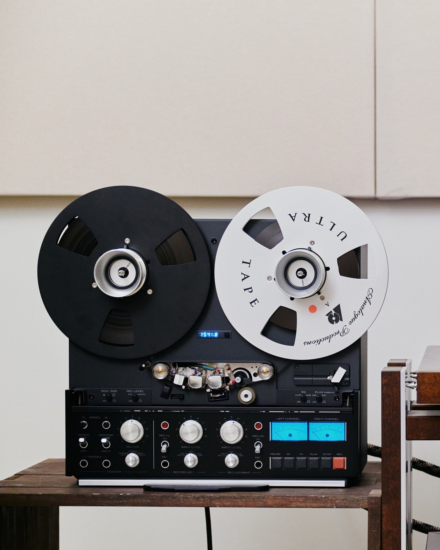 A vintage black reel-to-reel tape recorder with two large spools, one black and one white, sitting on a wooden table against a light beige wall.