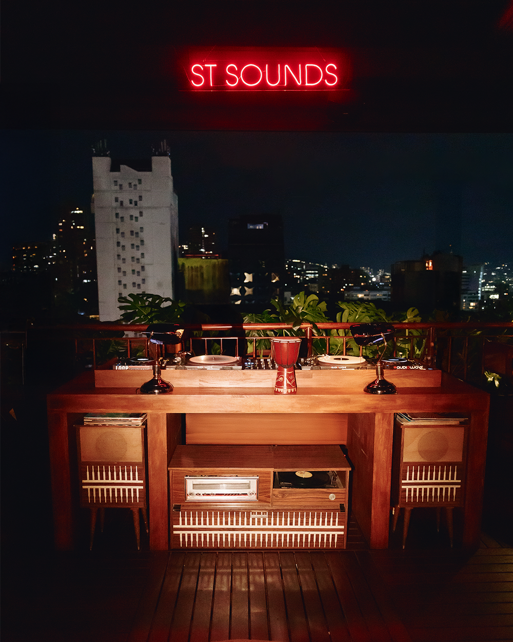 A wooden bar or dining table with chairs and wine glasses, set on a rooftop at night with a city skyline and a red neon sign reading 'ST SOUNDS' in the background.