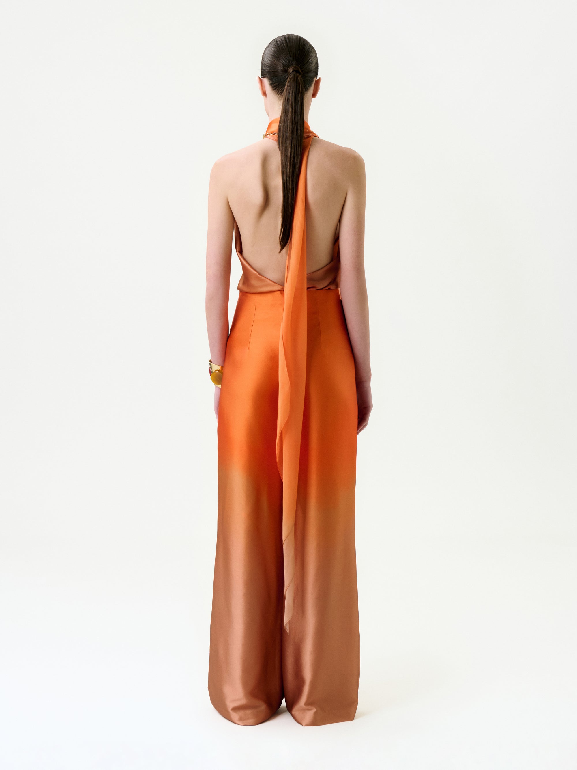 A woman with straight brown hair in a low ponytail is shown from behind, wearing the Andie Pant in orange Italian fabric, featuring a halter neck, long sash, wide legs, and a gold bracelet on her right wrist.