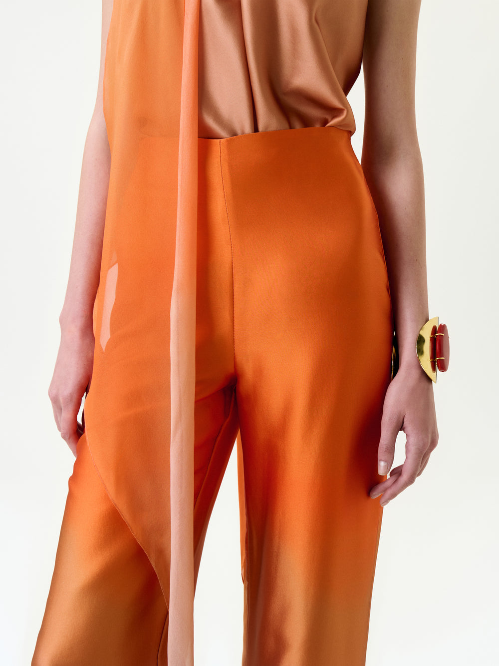 A person wears the Andie Pant in shiny orange Italian fabric with a peach top featuring a long scarf detail, accessorized by a chunky gold and red bracelet, all set against a plain white background.