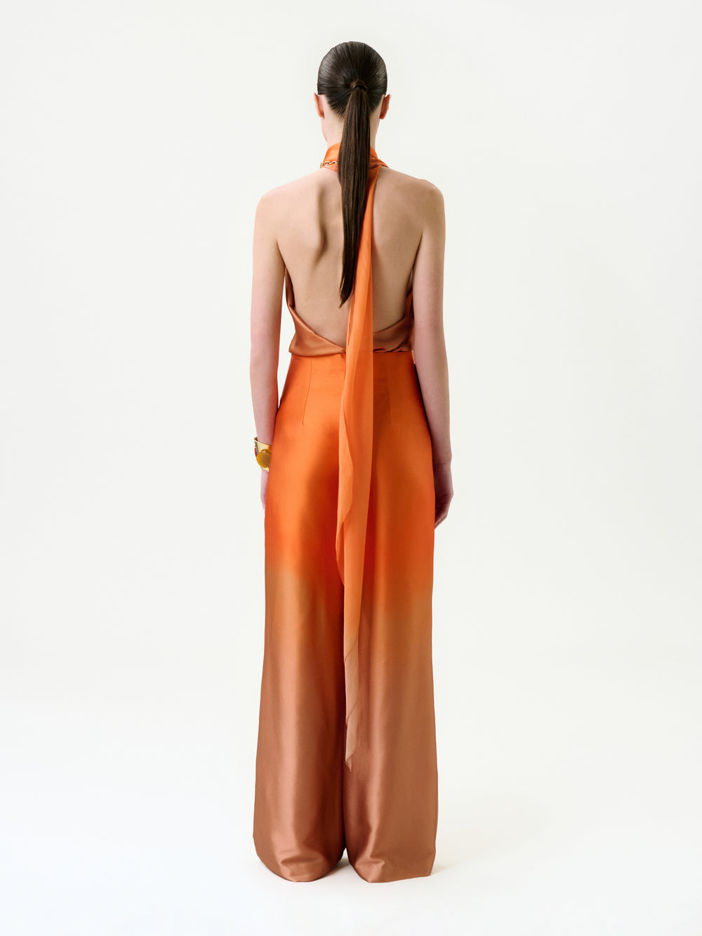 A woman with straight brown hair in a low ponytail wears the Velia Blouse, crafted from luxurious Italian fabric in an orange ombre hue, standing against a plain white background and facing away from the camera.