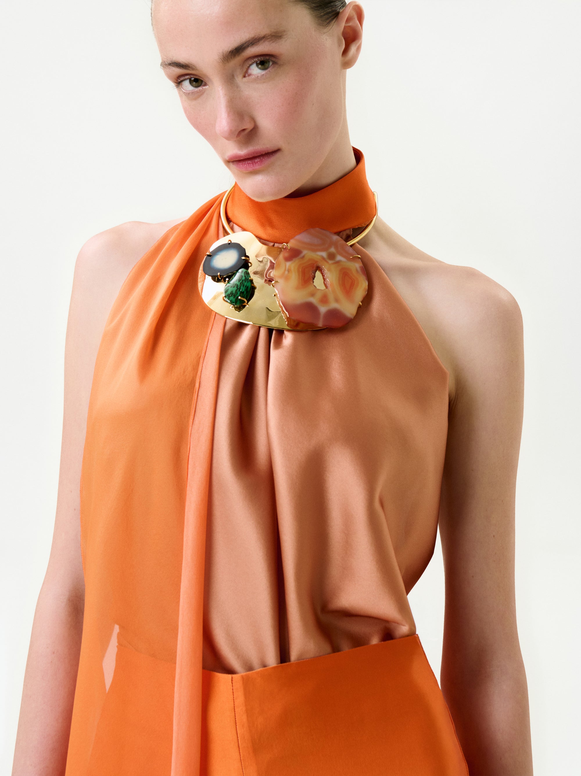 A woman with fair skin and pulled-back light hair wears the Velia Blouse—an orange, sleeveless, asymmetrical silk top—paired with a bold necklace of gold, black, green, and peach accents, posing against a plain white background.