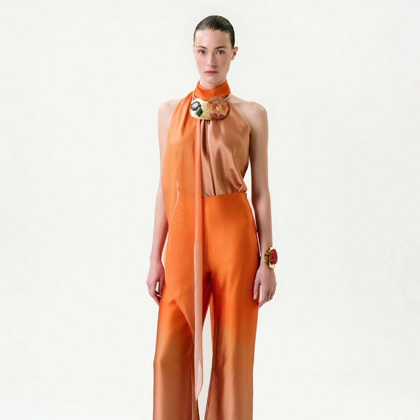 A woman stands against a white background wearing the Velia Blouse—an elegant sleeveless orange top with a high neck and sheer scarf detail, crafted from Italian fabric. She has slicked-back hair with a bold bracelet and brooch.