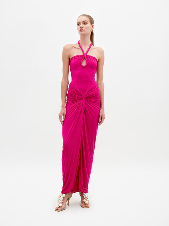 A woman stands in the Tori Dress Fuchsia, featuring a bright pink halter neck design with ruched detailing, paired with statement beige heels on a plain white background. This stylish ensemble is available for pre-order until February 15th, 2025.