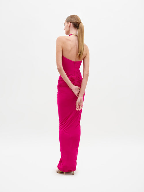 A woman stands in the Tori Dress Fuchsia, featuring a bright pink halter neck design with ruched detailing, paired with statement beige heels on a plain white background. This stylish ensemble is available for pre-order until February 15th, 2025.