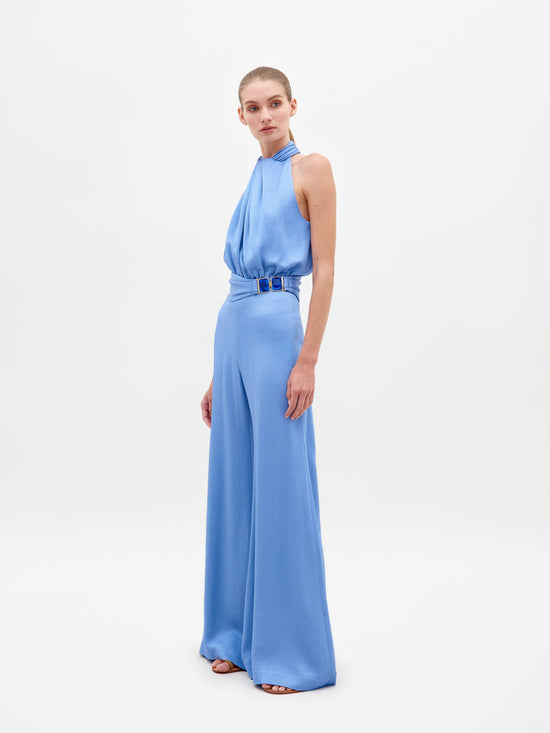 A person wearing the Siv Jumpsuit Periwinkle, featuring a sleeveless, light blue, halterneck design with wide-leg pants and a belted waist, stands against a plain white background. Pre-order now to ensure it ships by February 15th, 2025.