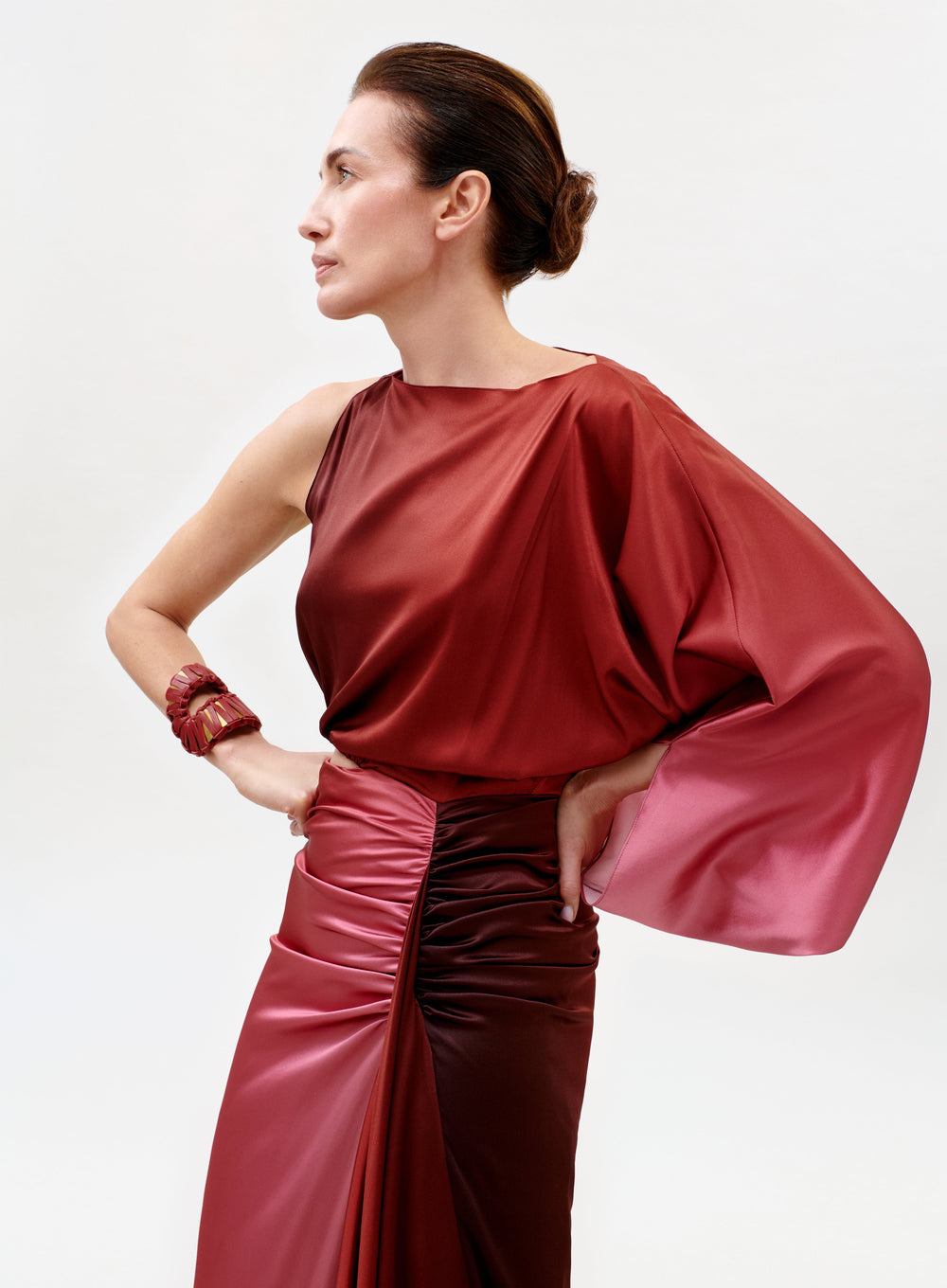 A woman in profile wears the Elimar Dress, a shiny, one-sleeved red satin gown with a gathered waist. Her hair is styled in a sleek updo, and she poses with one hand on her hip, accessorized with a matching red bracelet against a light background.