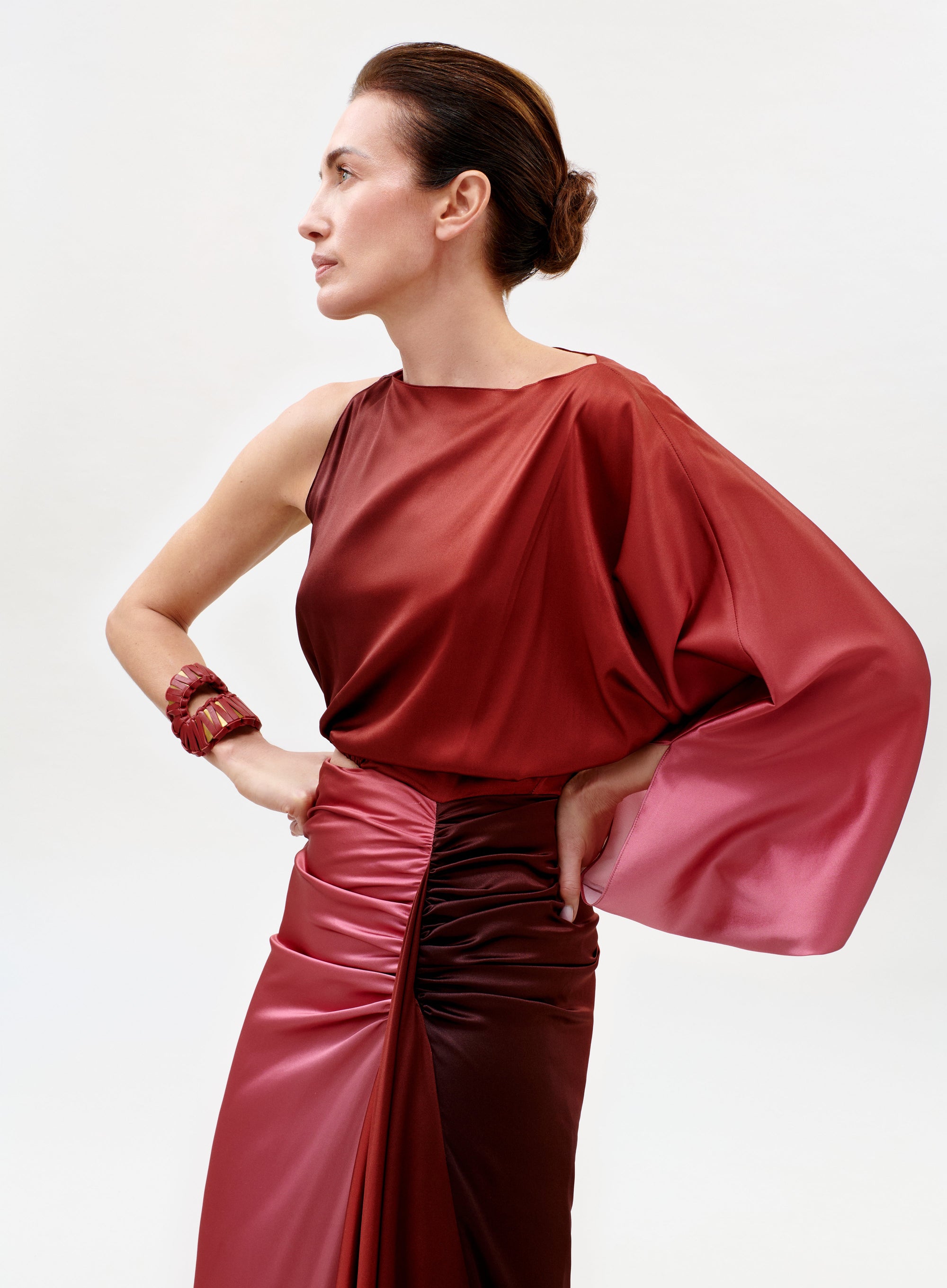 A woman in profile wears the Elimar Dress, a shiny, one-sleeved red satin gown with a gathered waist. Her hair is styled in a sleek updo, and she poses with one hand on her hip, accessorized with a matching red bracelet against a light background.