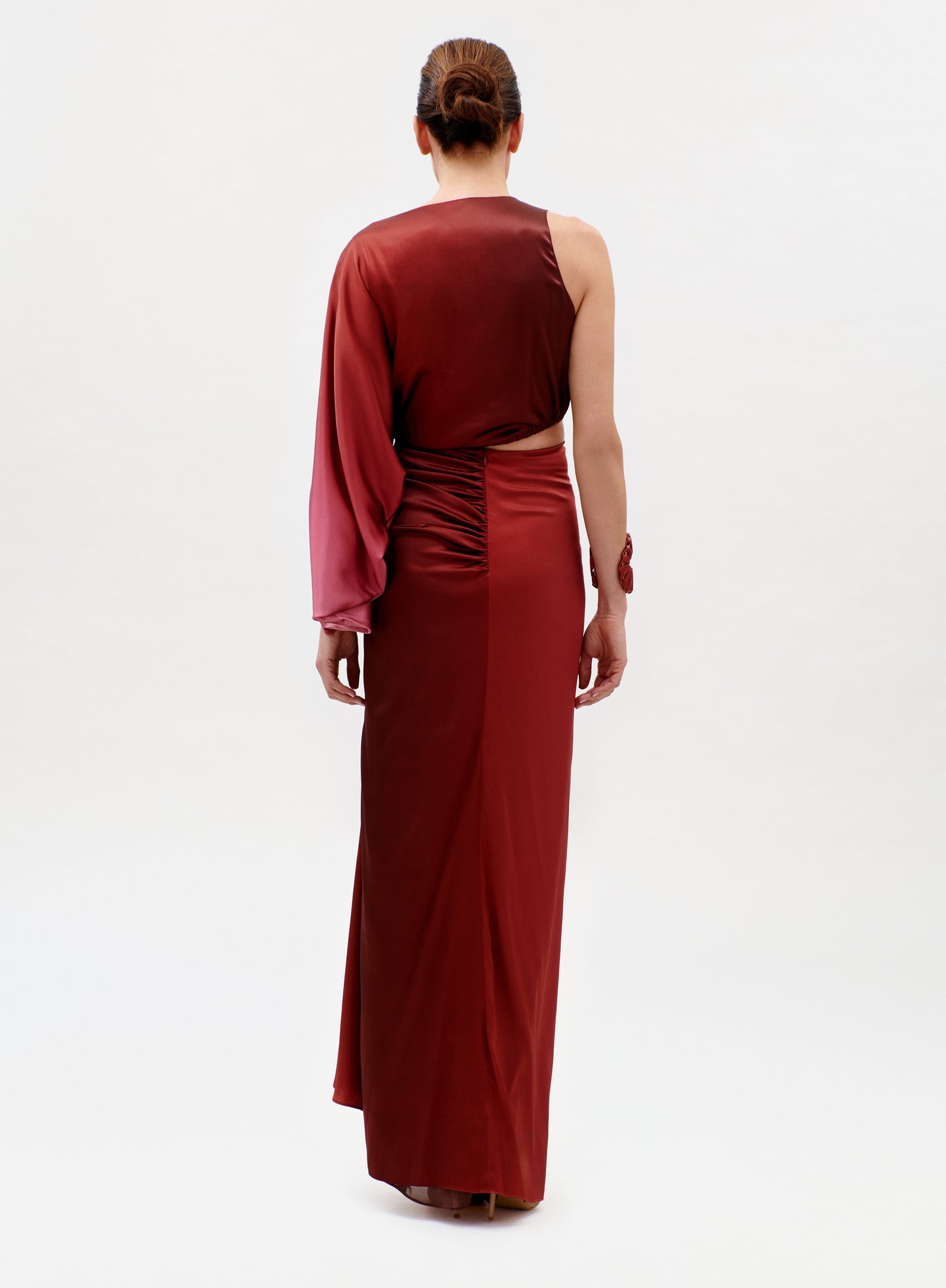 A woman with her hair in a neat bun stands facing away, wearing the Elimar Dress—a floor-length dark red gown with a single long sleeve on the left arm—against a plain light-colored background.