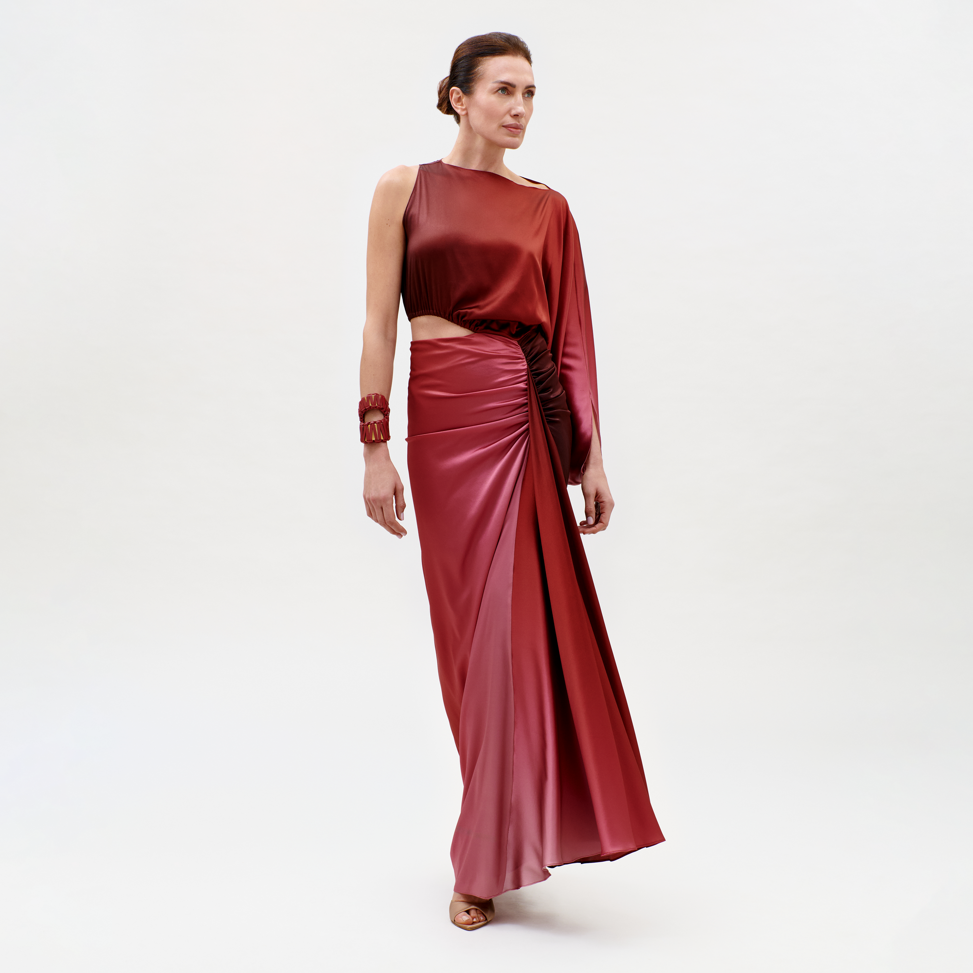 A woman wears the Elimar Dress—a shiny dark red gown with one long sleeve, an asymmetrical waist cutout, and a draped skirt. Her hair is pulled back, and she accessorizes with a matching bracelet.