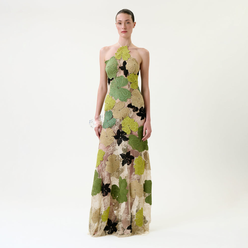 A person stands against a white background wearing the Xenia Dress—a floor-length, sleeveless gown featuring large green, yellow, and black floral patterns on sheer fabric.