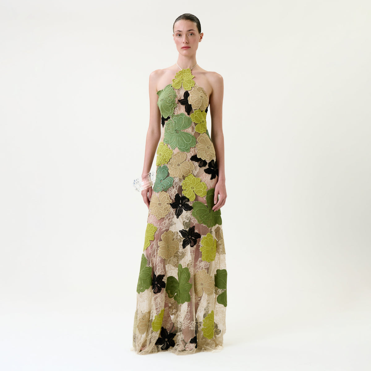 A person stands against a white background wearing the Xenia Dress—a floor-length, sleeveless gown featuring large green, yellow, and black floral patterns on sheer fabric.