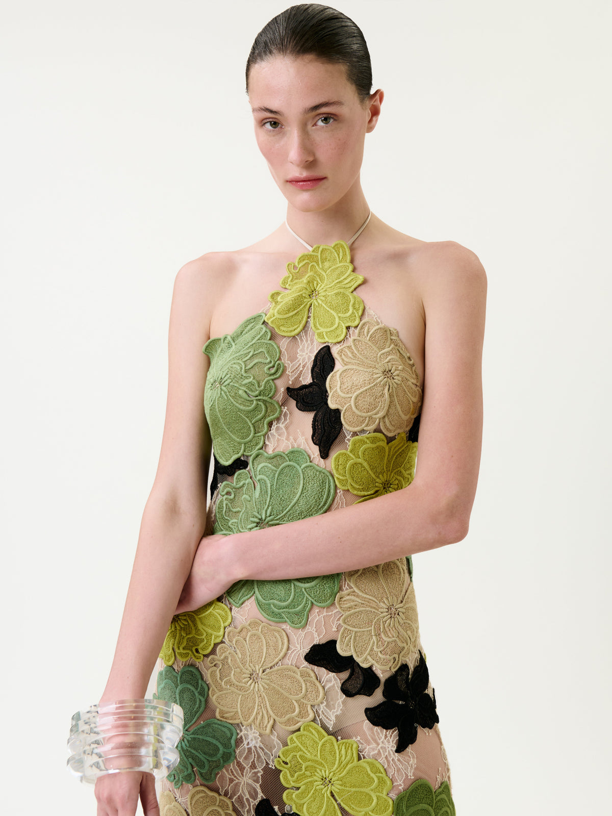 Woman with slicked-back hair wears the Xenia Dress—a sheer design adorned with large green, yellow, and black floral appliqués—paired with a thin necklace and a chunky clear bracelet, standing against a plain light background.